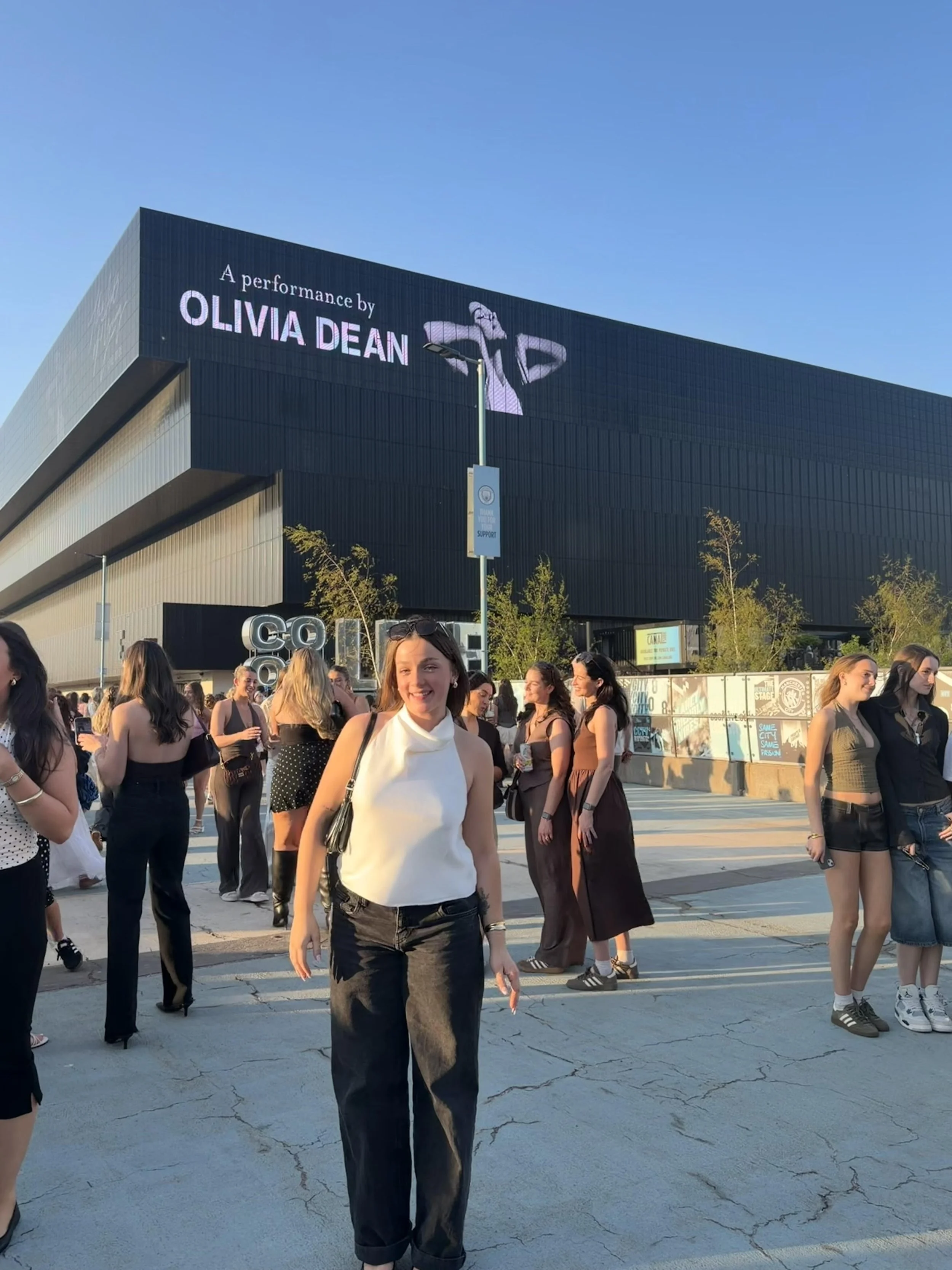 Group of people gathered outdoors in front of a building with a sign that reads 'A performance by Olivia Dean.' The sky is clear and the sun is setting.