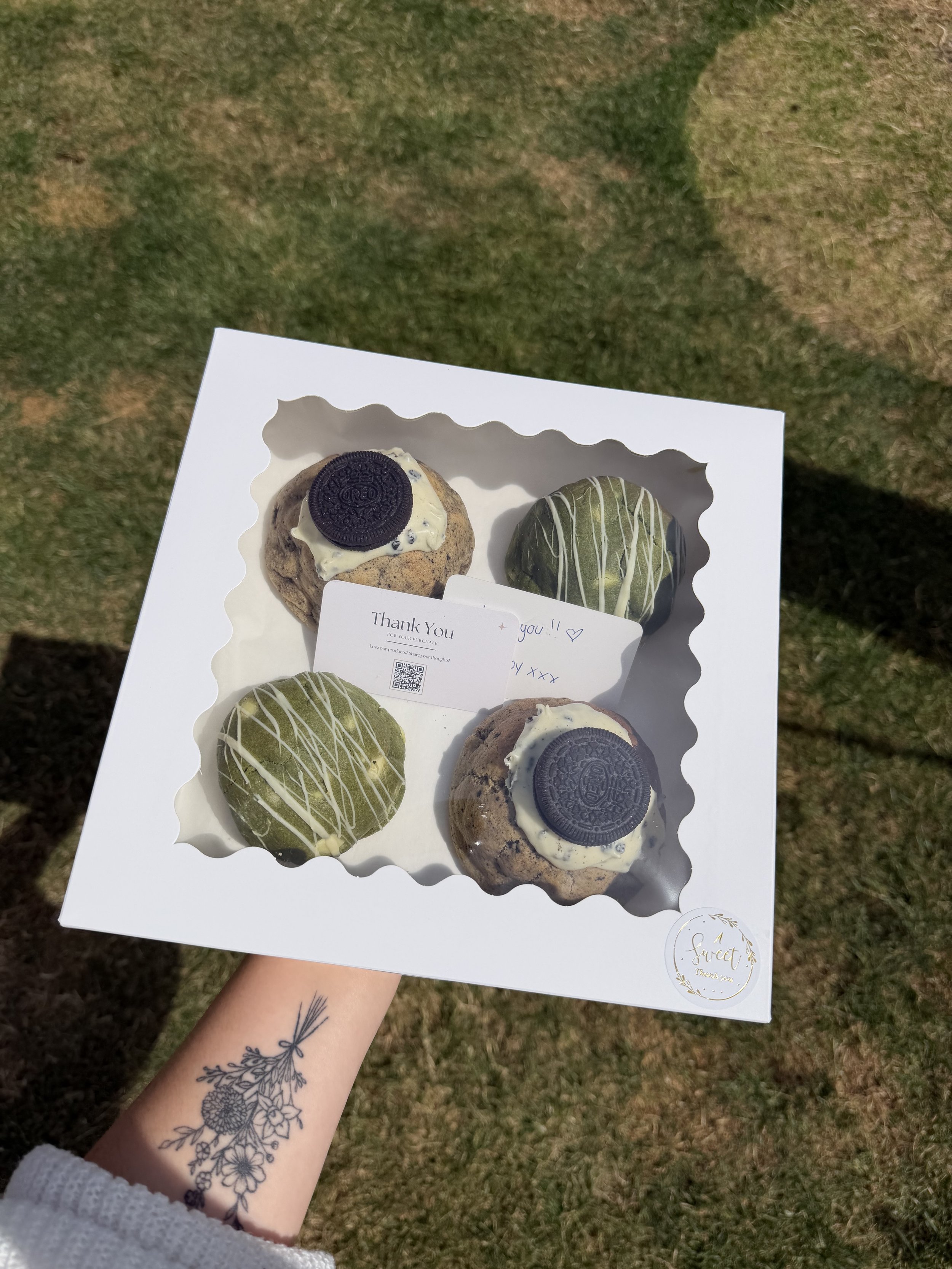 Person holding a box of four decorated cookies outdoors on grass, with a tattoo of flowers on their arm.