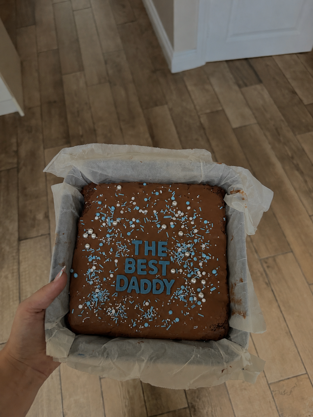 A hand holding a brownie slab decorated with blue and silver sprinkles, with blue letters spelling 'THE BEST DADDY' on top, in a baking pan lined with parchment paper.