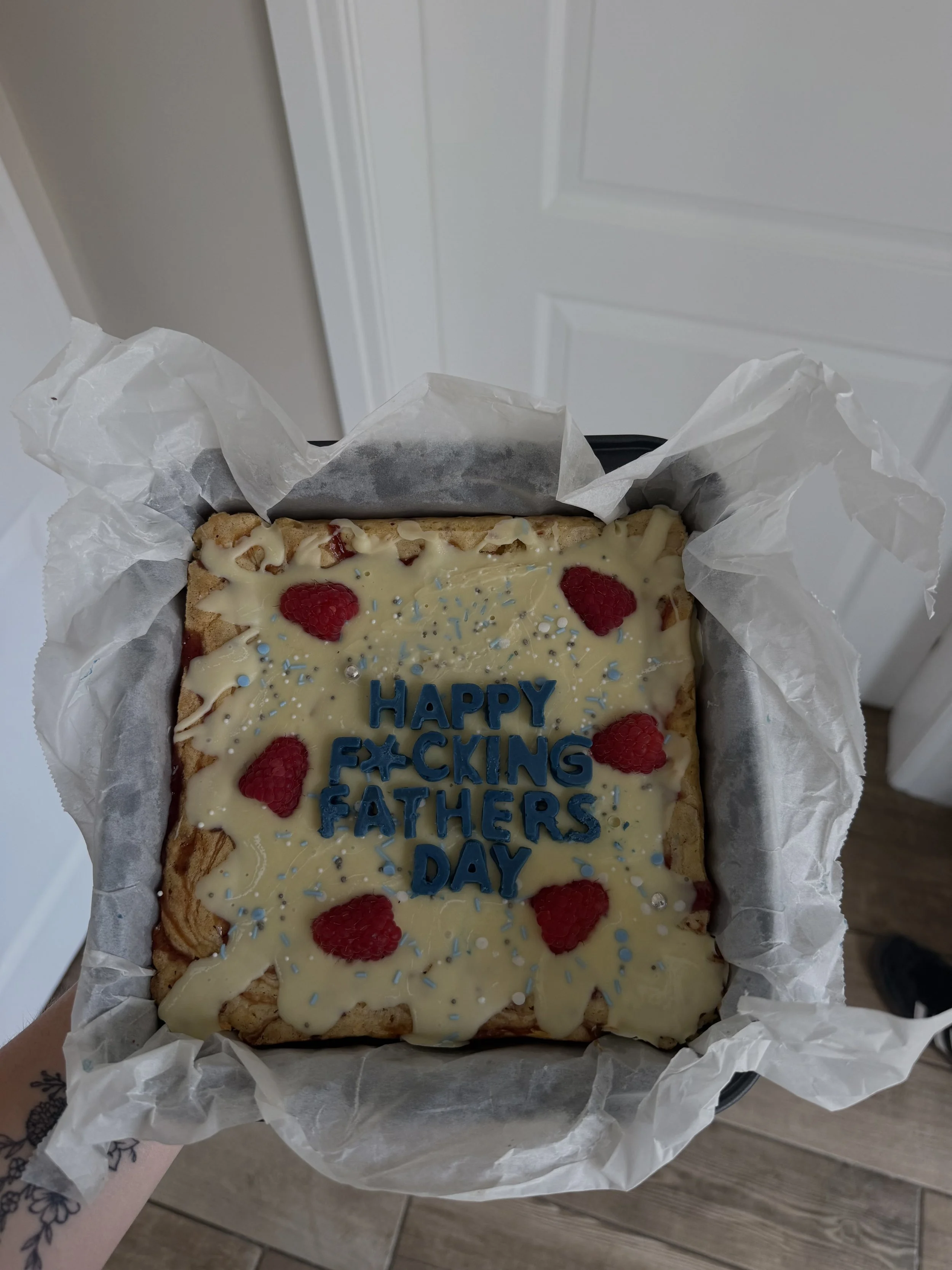A rectangular dessert with white chocolate, red raspberries, and blue sprinkles, inscribed with a message for Father's Day, with a partly torn white parchment paper lining. The message reads 'HAPPY F*CKING FATHERS' DAY' with some letters obscured.