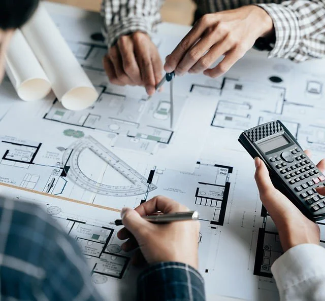 Two people are working on architectural plans with blueprints, a calculator, and a pencil on a table.