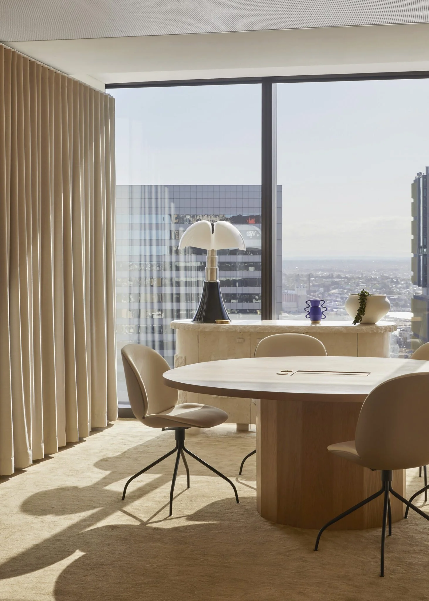 Modern interior of a high-rise office with a round table, beige chairs, decorative vases, a lamp, large windows showing a cityscape, and beige curtains.