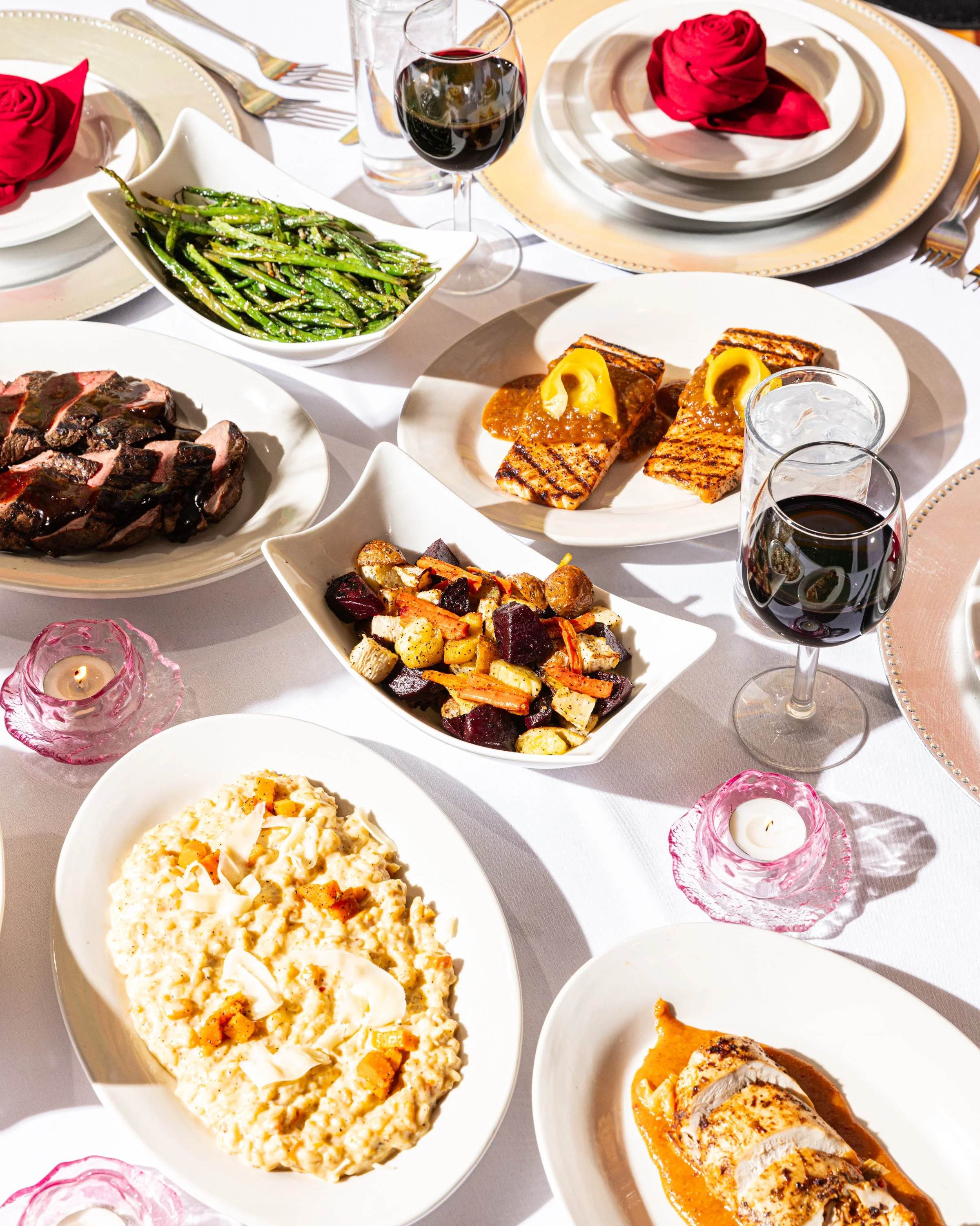 A formal dining table with white tablecloth, featuring plates with food, red roses, wine glasses filled with red wine, a glass of water, and pink candles in glass holders.