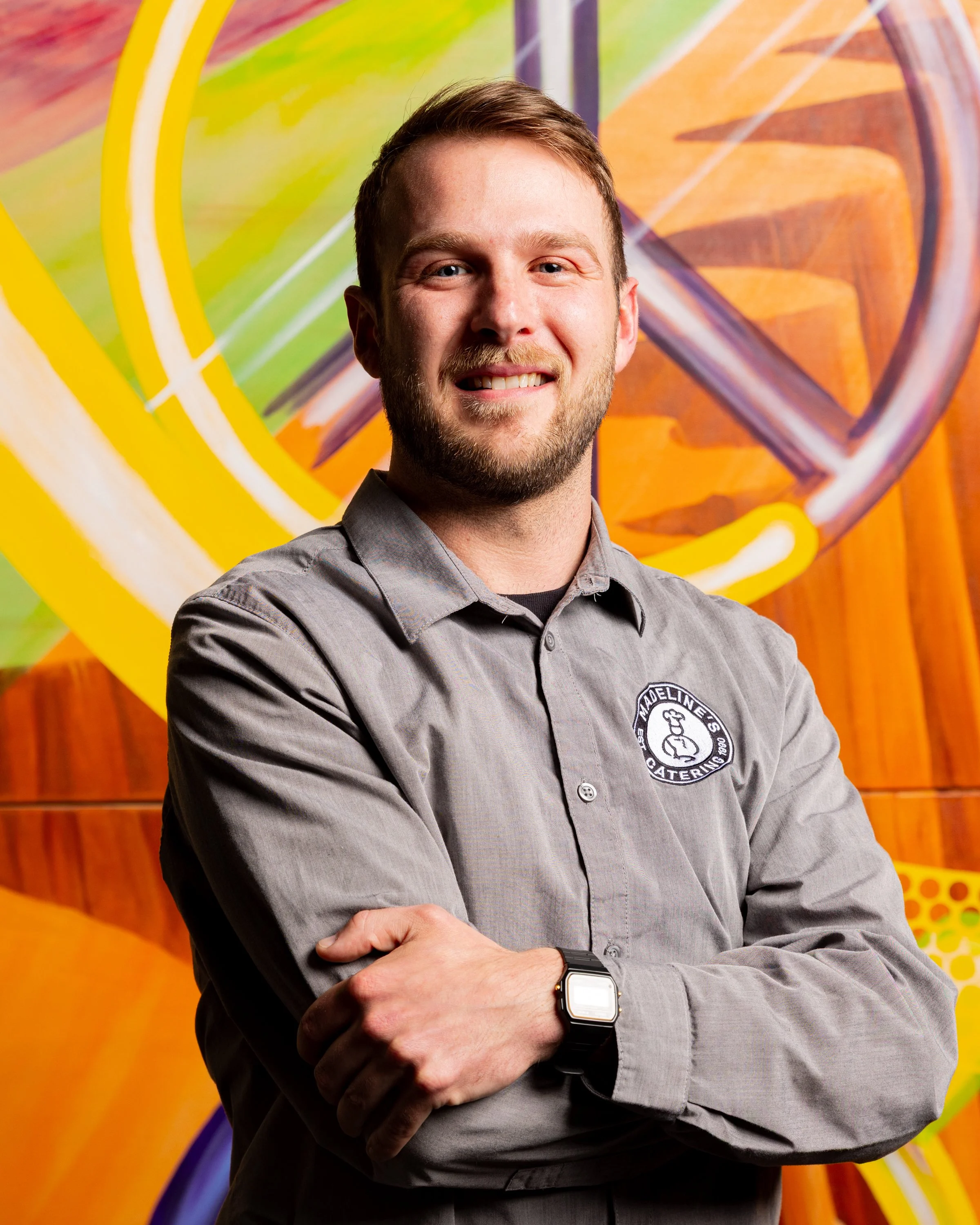 A young man with light brown hair and a beard smiling with crossed arms, wearing a gray button-up shirt and a black smartwatch, standing in front of a colorful, abstract mural with geometric shapes and bold colors.