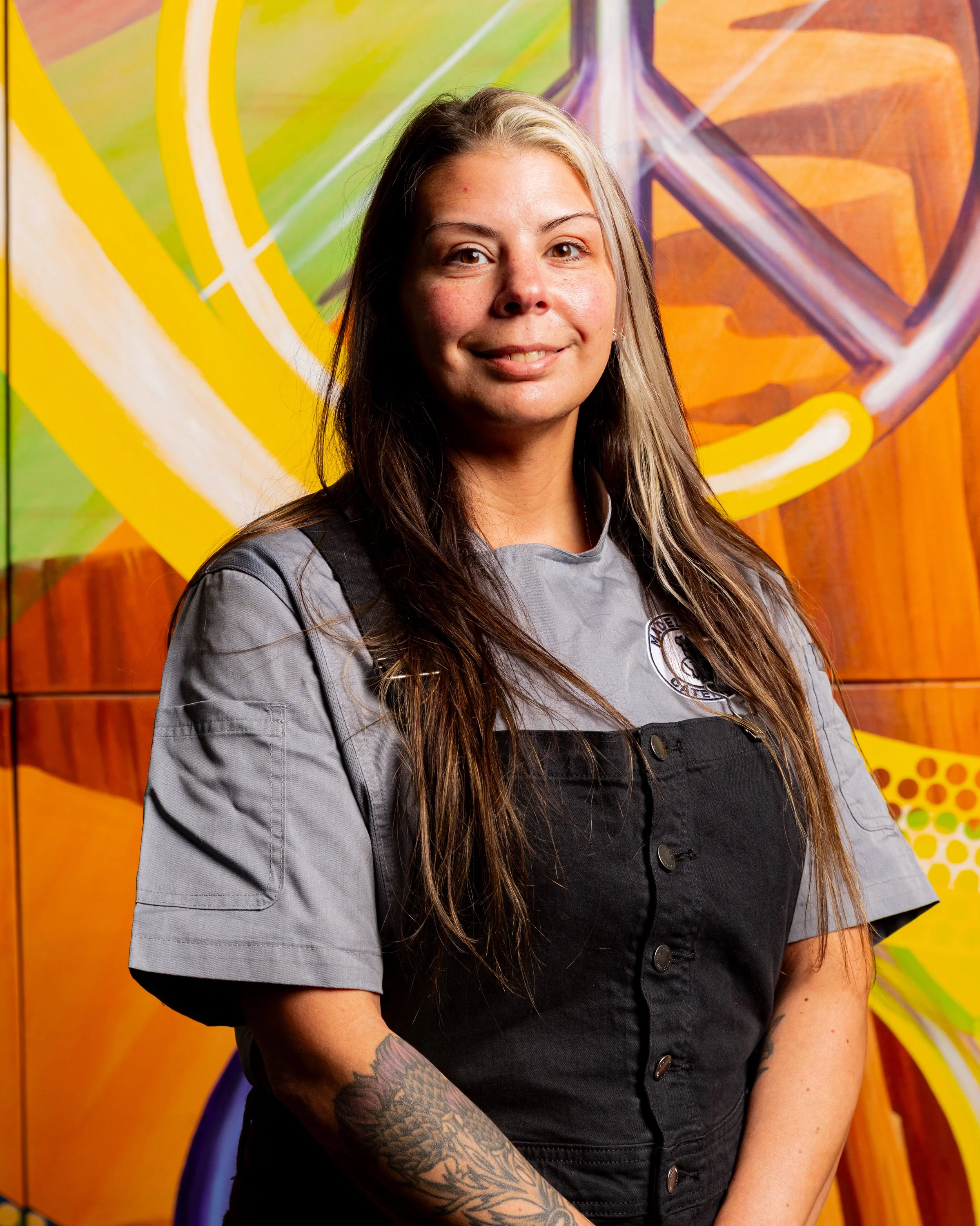 Portrait of a woman with long brown hair, tattoos on her arm, wearing a gray short-sleeved top and black vest, standing in front of a colorful graffiti background.