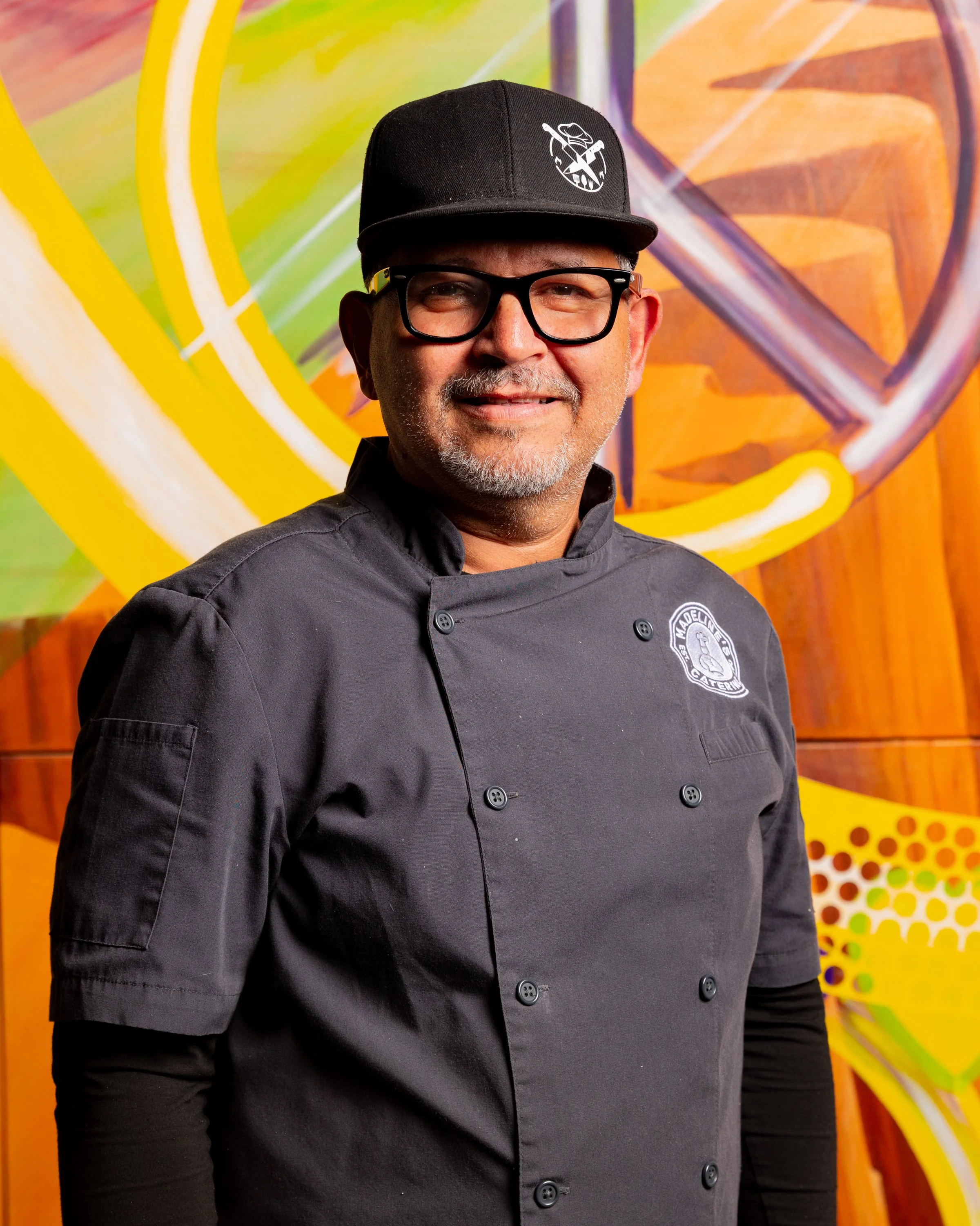 A man wearing glasses and a black chef's jacket with a logo, standing in front of a colorful abstract background.