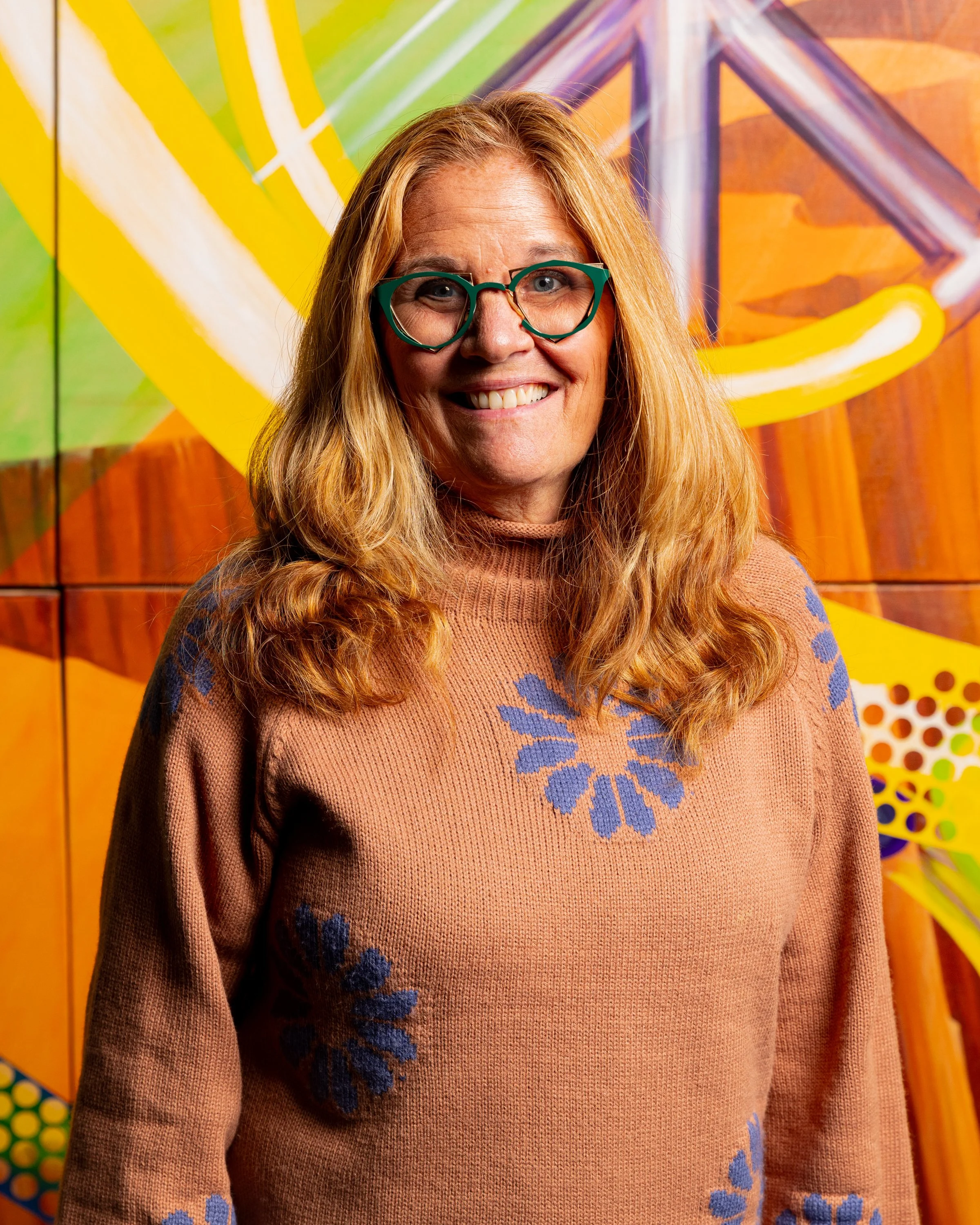 A smiling woman with red hair and glasses standing in front of colorful, abstract wall art.