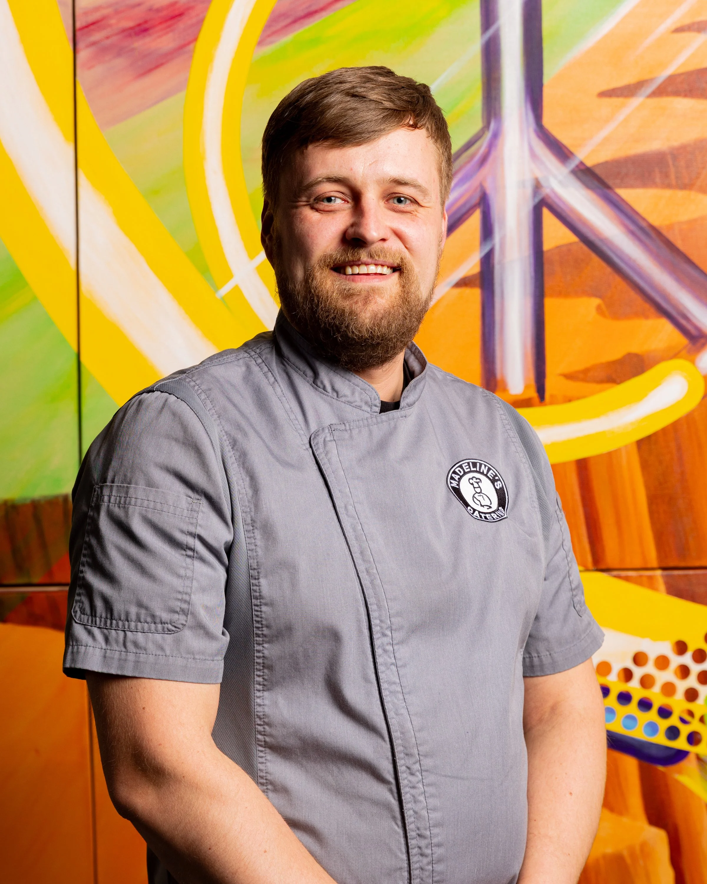 A man with short brown hair, a full beard, and a gray chef's jacket smiling in front of a colorful abstract mural featuring bright colors and geometric shapes.