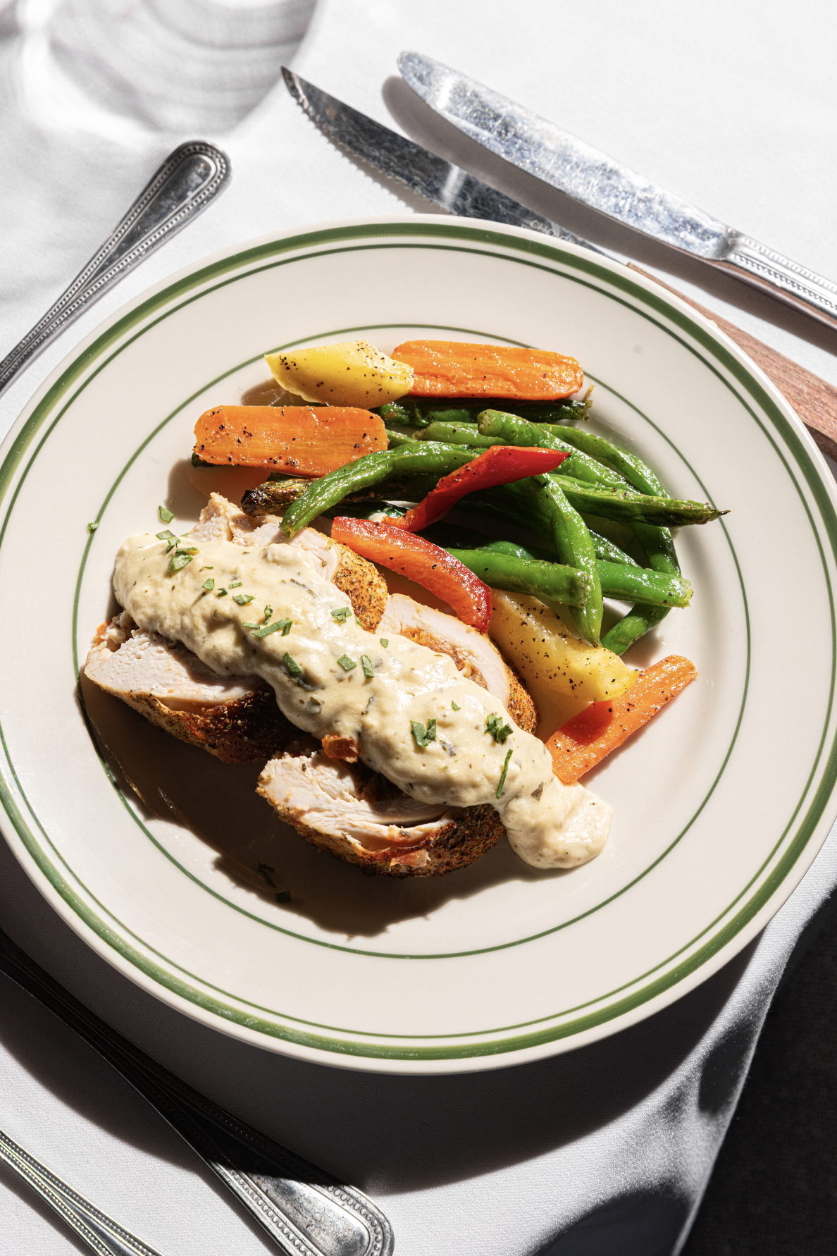 Plate of roasted turkey with gravy, mixed roasted vegetables including carrots, green beans, and red bell peppers, on a white dish with green rings, set on a white tablecloth.