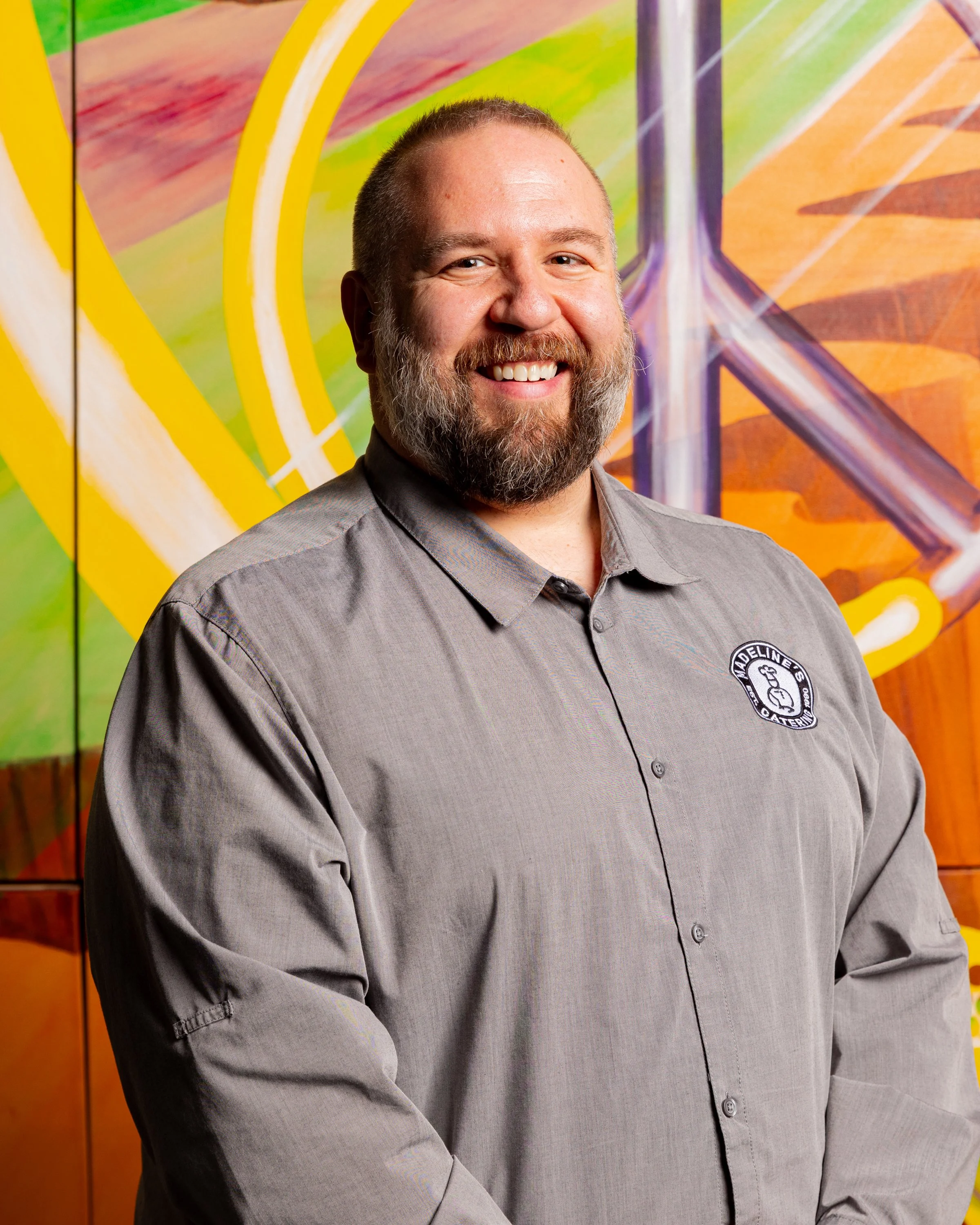 A man with a beard and short hair smiling, wearing a gray shirt with a logo on the chest, standing in front of a colorful abstract mural.