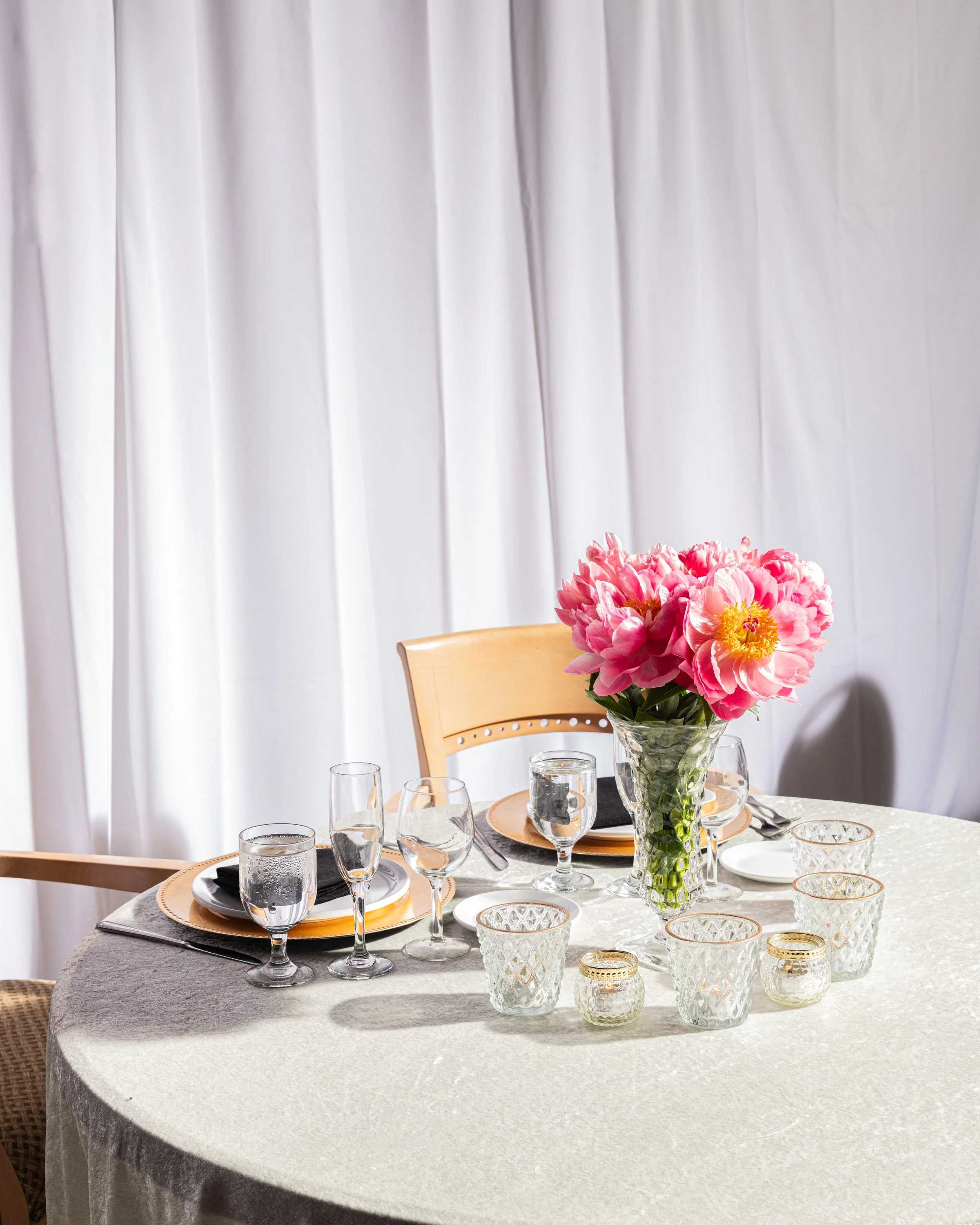 Elegant dinner table set with glassware, plates, and a floral centerpiece of pink flowers in a glass vase, with a white cloth backdrop.
