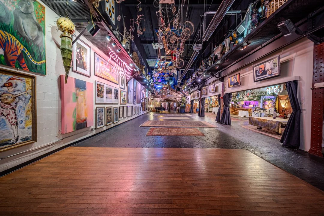 Empty event space with round tables covered in white tablecloths, black chairs, colorful artwork on the walls, and artistic installations hanging from the ceiling.