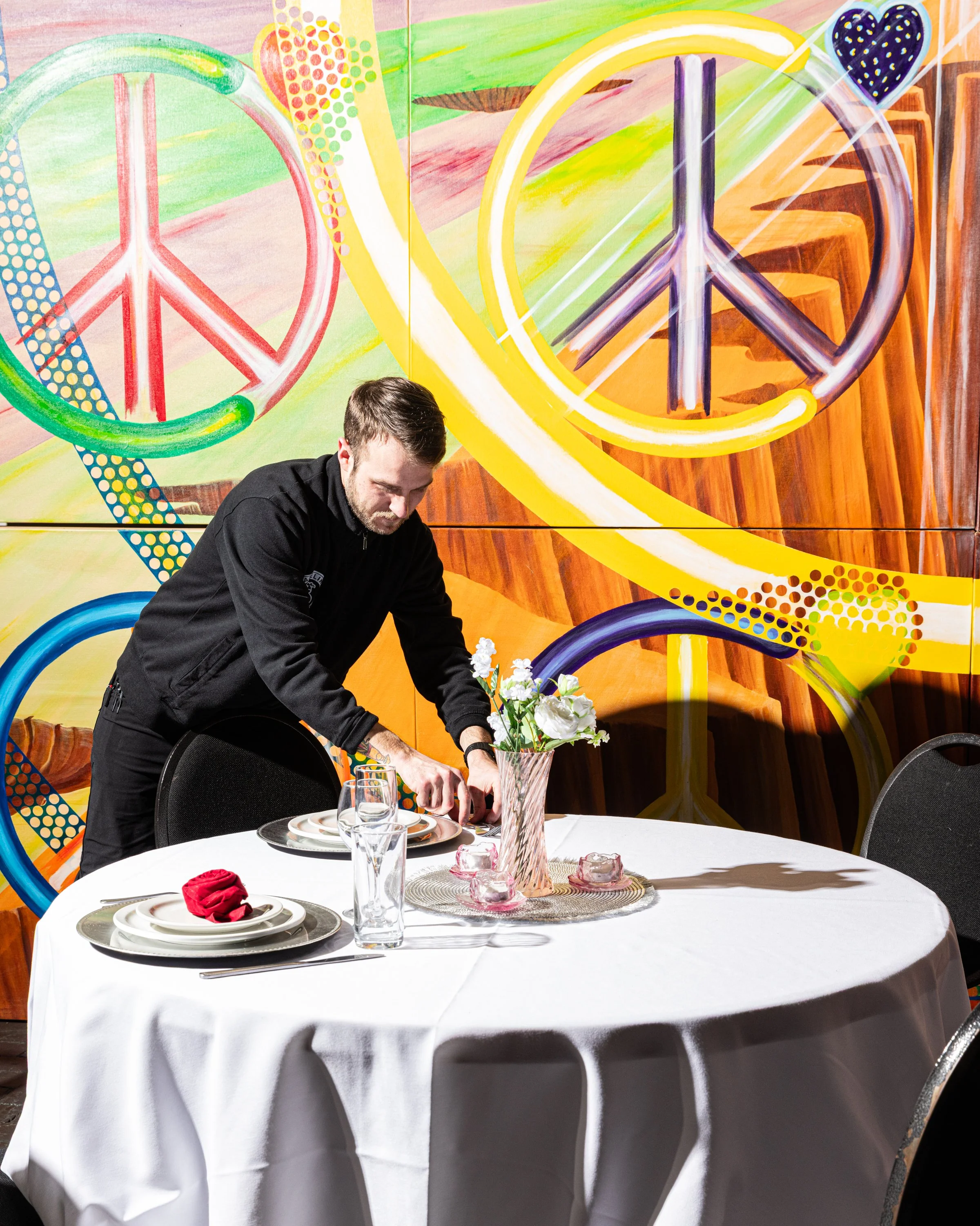 A man preparing a table set with plates, glasses, napkins, and a pink vase with white flowers in front of a colorful graffiti mural featuring peace signs and vibrant colors.