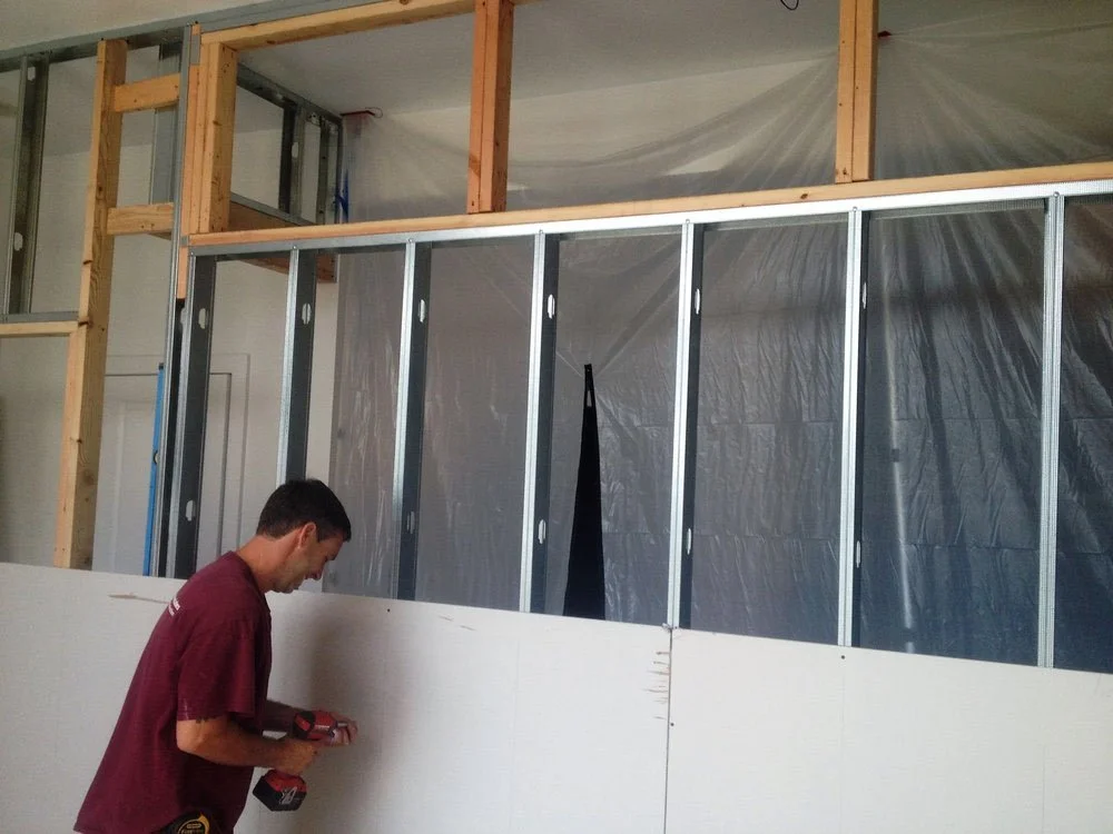 A man working on a construction site, installing insulation or drywall in a room with exposed wooden framing and metal studs.