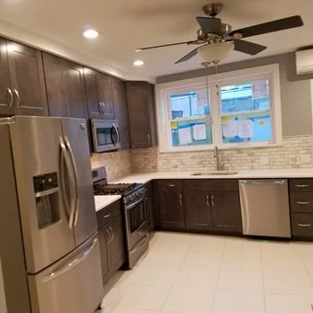 Modern kitchen with dark wood cabinets, stainless steel appliances including refrigerator, oven, microwave, and dishwasher, a white tiled backsplash, and a ceiling fan with lights.