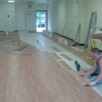 A person installing laminate flooring in a large room, with tools and flooring materials scattered around.