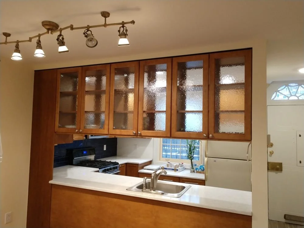 Kitchen with wooden cabinets, a white countertop, a sink, a stove, a window with blinds, and a plant. Overhead track lighting with multiple spotlights.