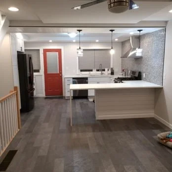 Modern kitchen with white cabinets, a red door, and pendant lights hanging over a kitchen island, with a gray wood floor.