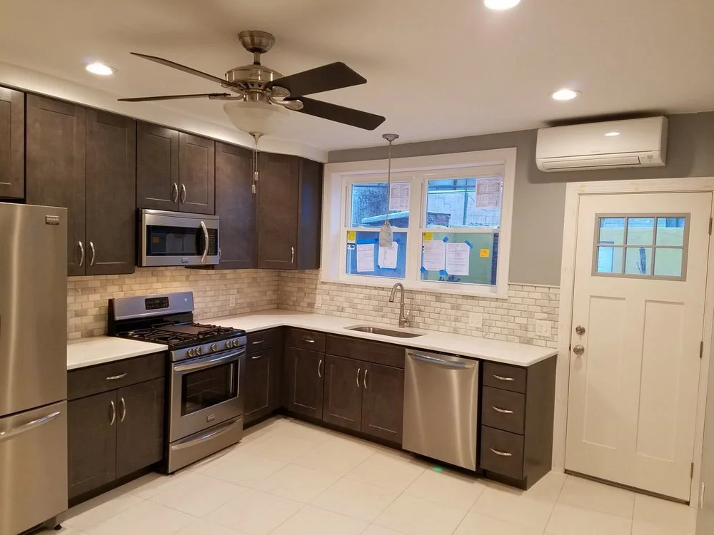 Modern kitchen with dark cabinets, stainless steel appliances, white countertops, brick backsplash, window, ceiling fan, and wall-mounted air conditioner.