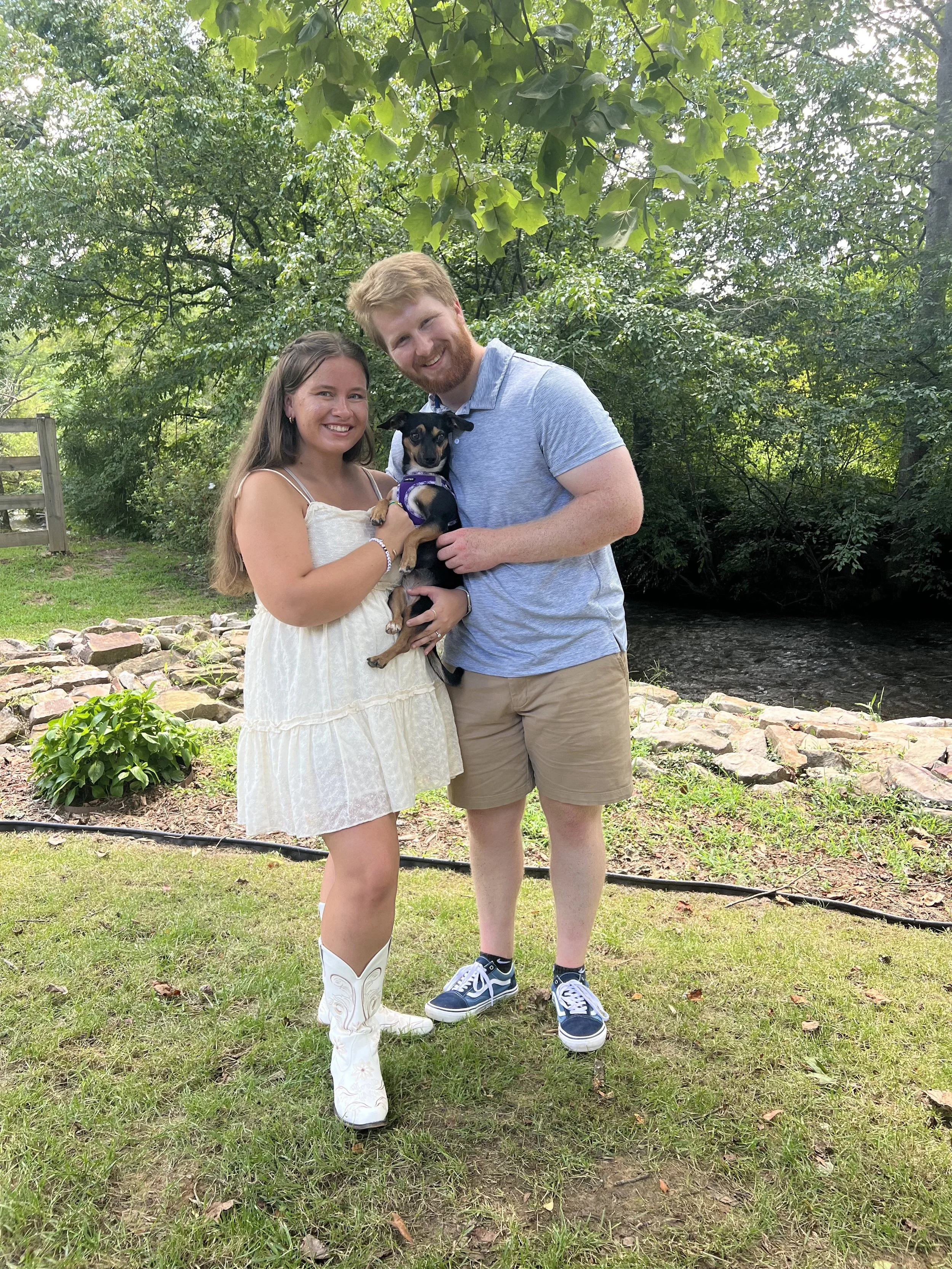A smiling couple, a woman and a man, holding a small dog in a park with lush green trees and grass, rocks, and a wooden fence in the background.