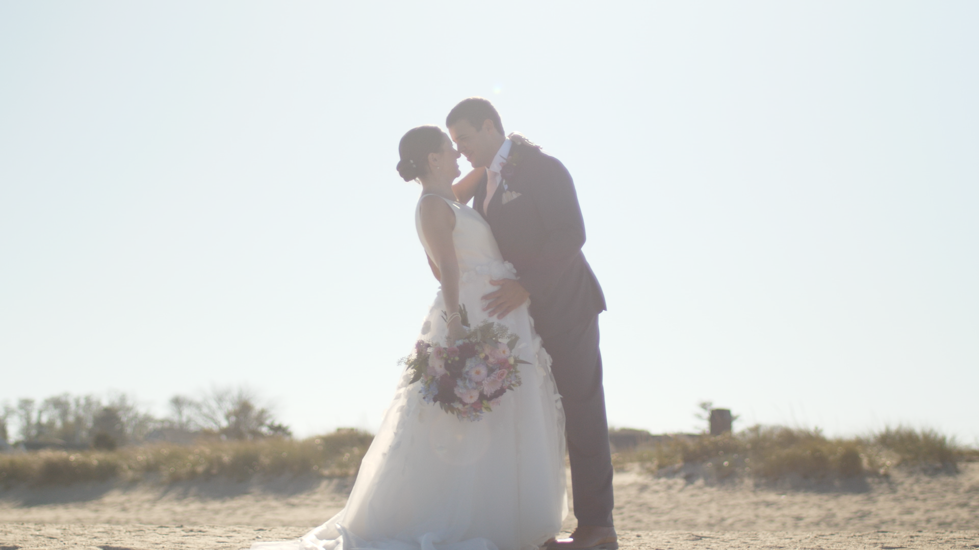 A bride and groom embracing outdoors, with the bride holding a bouquet of flowers, backlit by the sun against a clear sky.