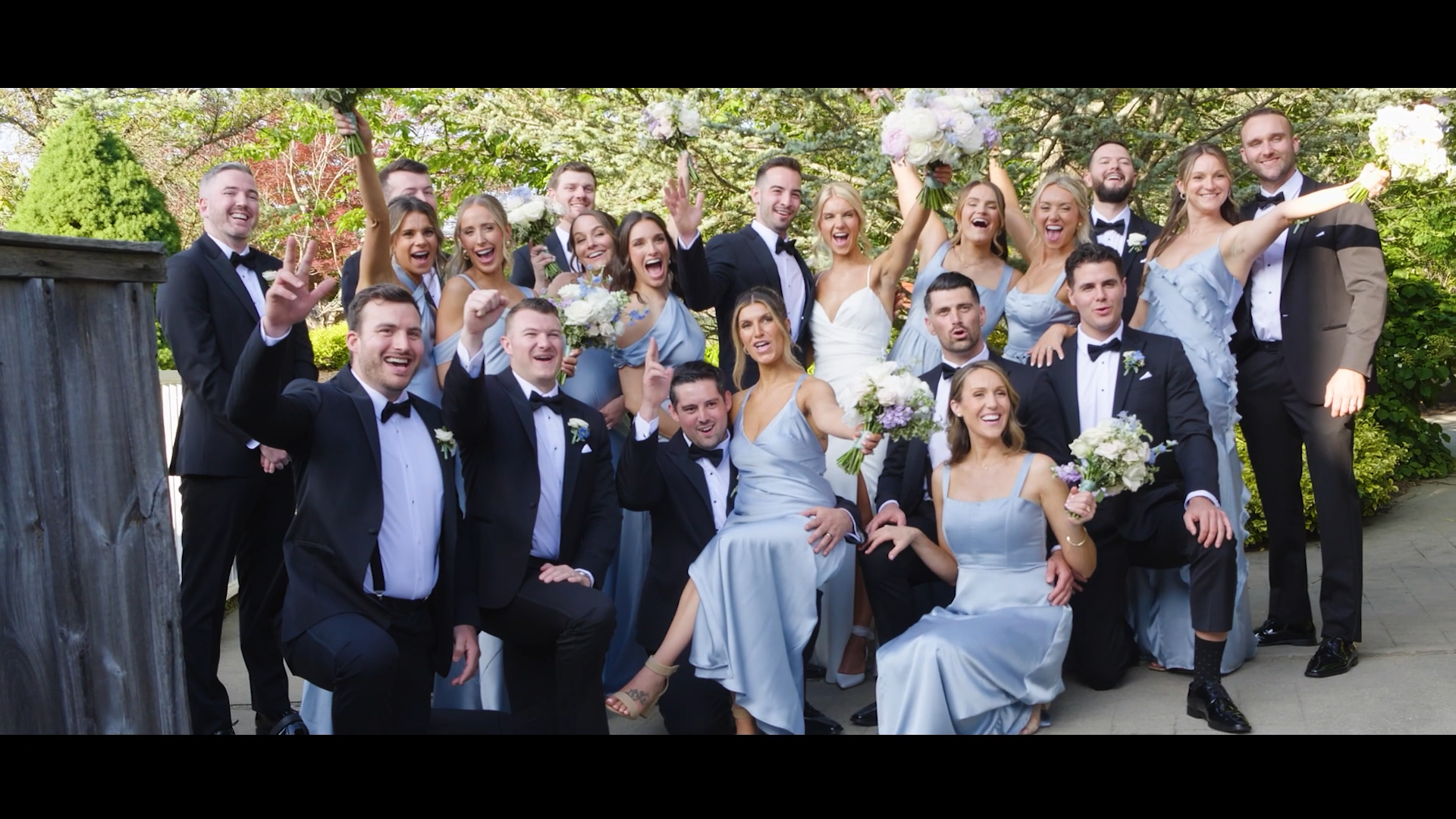 A newlywed couple and their wedding party celebrating outdoors, with the bride in a white dress holding a bouquet, surrounded by bridesmaids in light blue dresses and groomsmen in tuxedos, all smiling and joyfully posing for the camera.