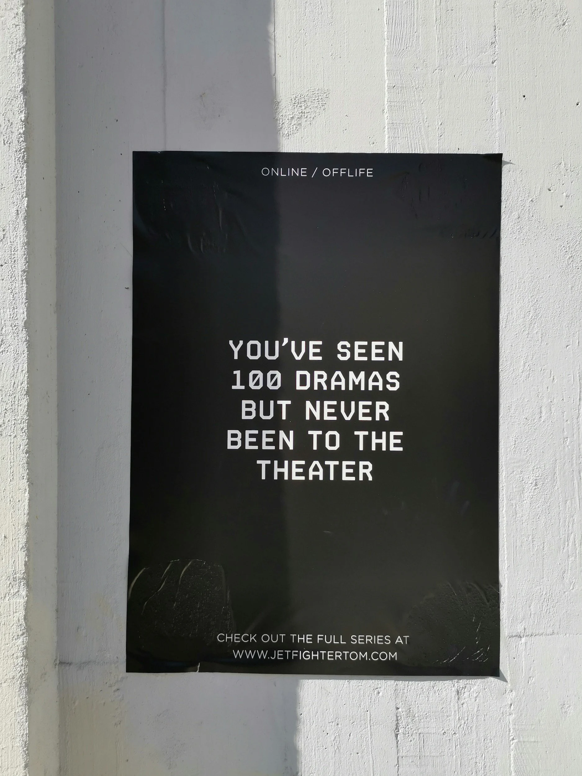 A black poster on a white wall with white text reading: 'Online / Offline. You've seen 100 dramas but never been to the theater. Check out the full series at www.jetfighertom.com'.