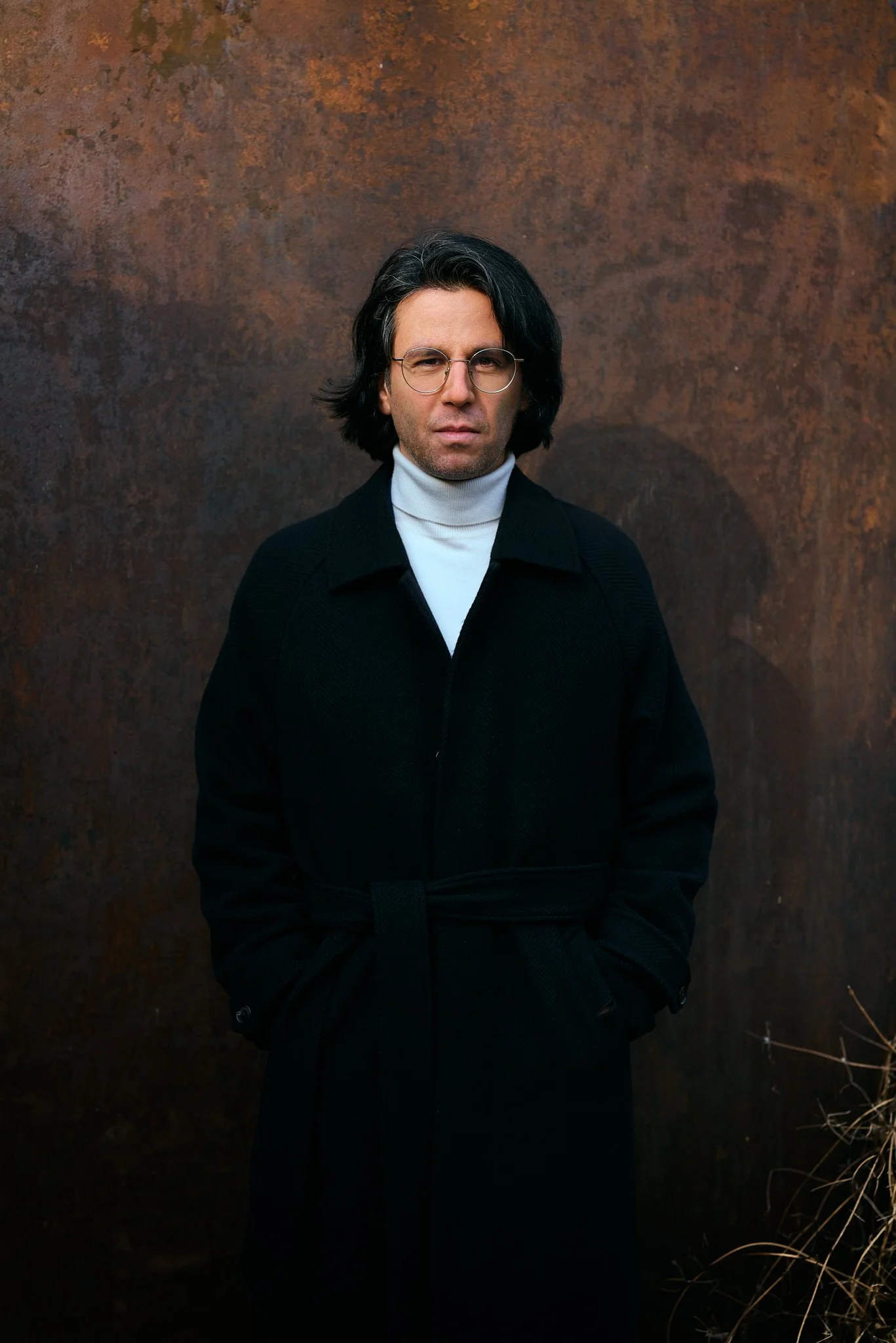 Portrait von Deniz Utlu. Ein Mann mit mittellangen schwarzen Haaren, trägt eine schwarze Jacke und eine weiße Turtleneck. Steht vor einer rostigen Metallwand im Freien.