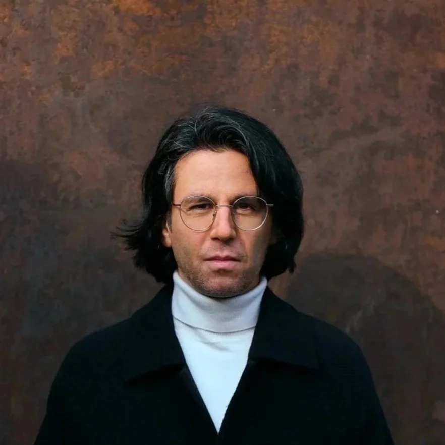 Porträt von Deniz Utlu. Eine Mann mit schwarzen Haaren, Brille und weißen Rollkragenpullover vor einer rostfarbenen Metallwand.