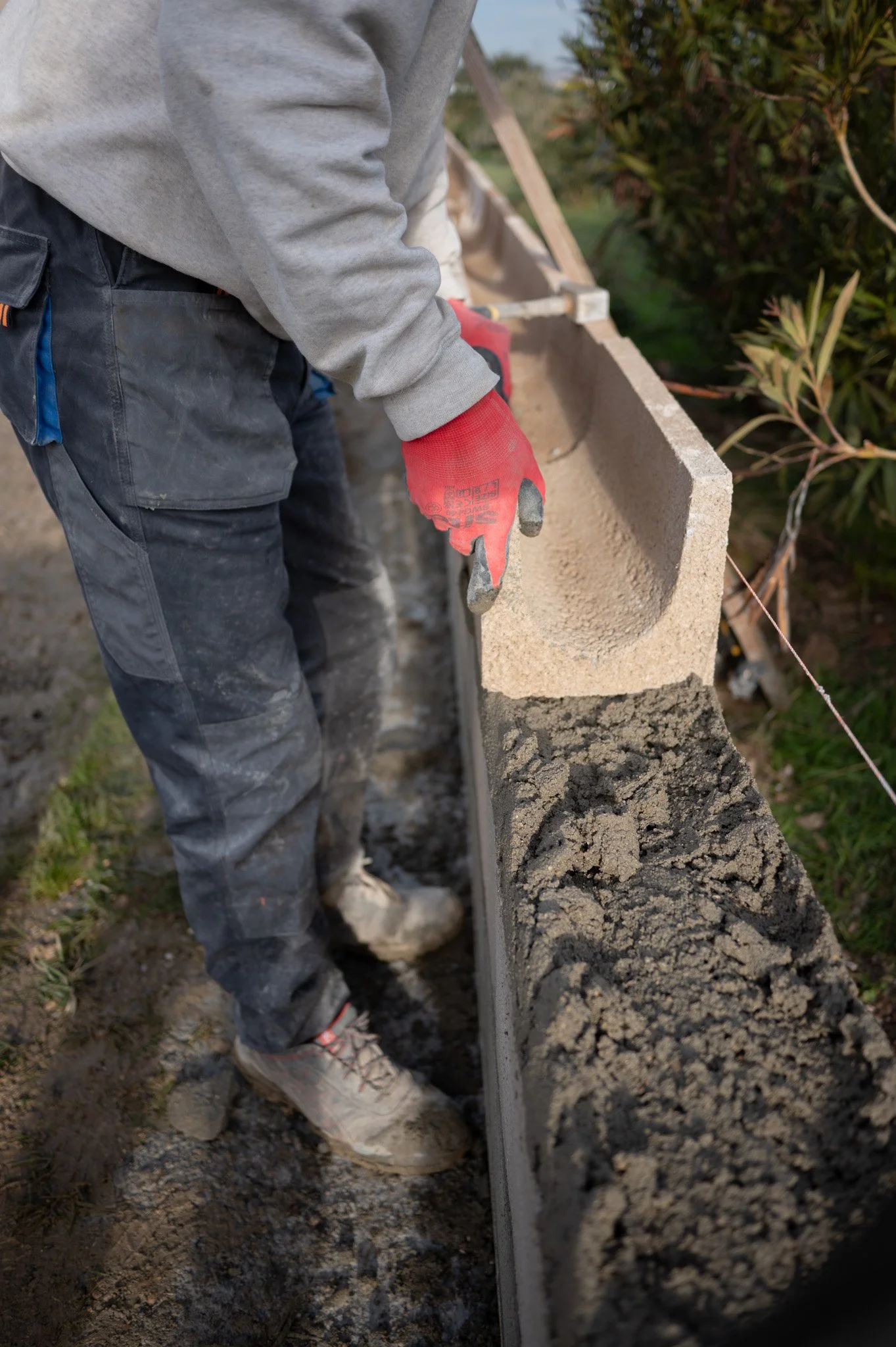 Un ouvrier en train de poser un mur en blocs en béton, portant des gants rouges, dans un environnement extérieur avec des arbres en arrière-plan.
