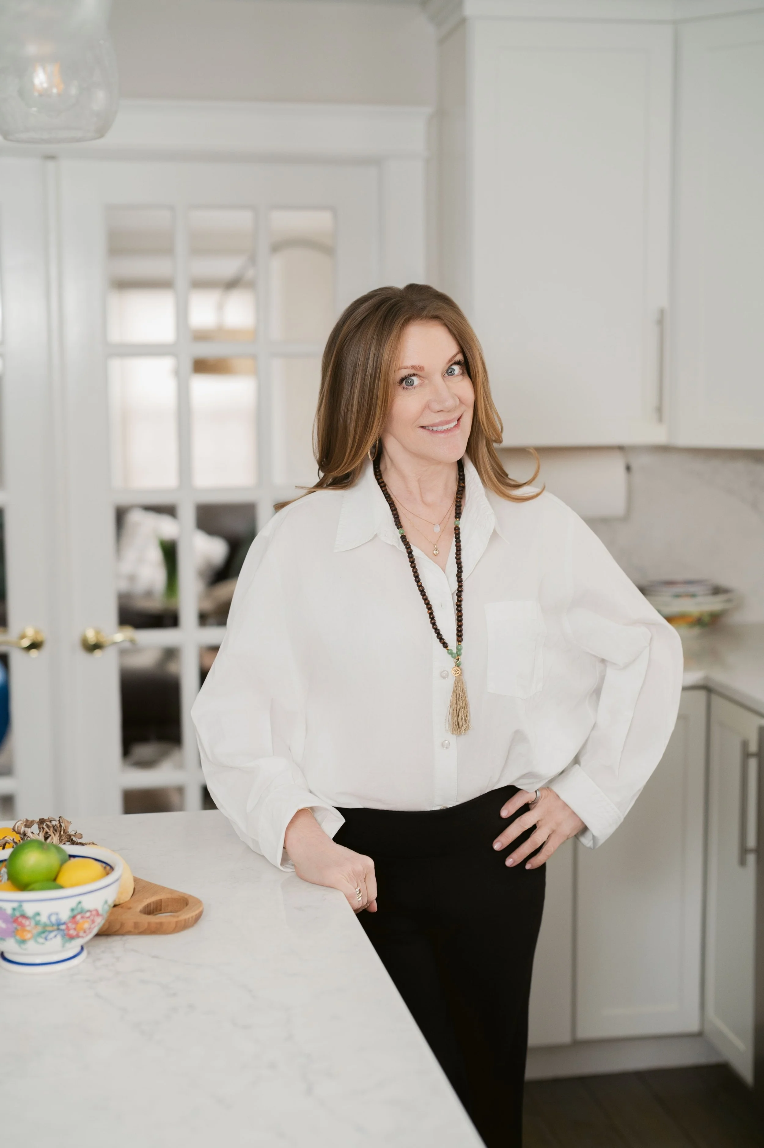 A woman with reddish-brown hair, wearing a white shirt and black pants, standing in a kitchen near a white countertop. There is a bowl of lemons and limes on the counter.