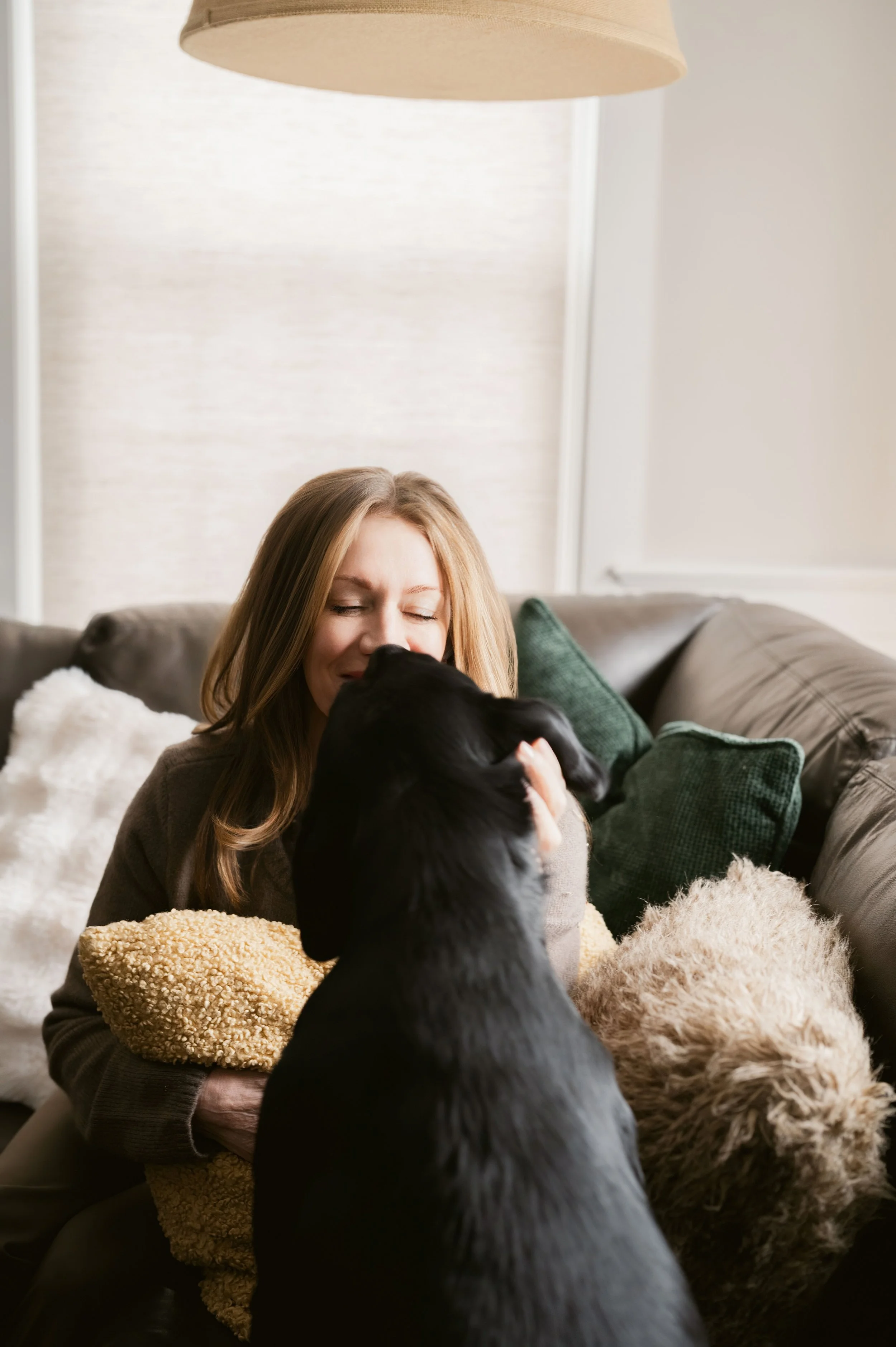 A woman with closed eyes happily hugging a black dog on a sofa, surrounded by fluffy pillows and cozy blankets in a living room.