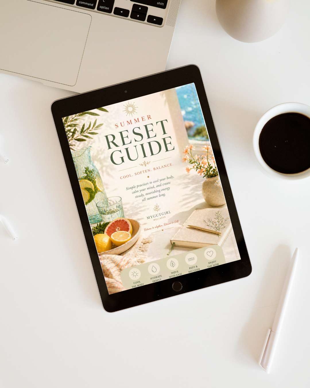 Tablet displaying a summer reset guide with images of citrus fruits, flowers, and a beach setting, surrounded by a cup of coffee, a laptop, a white pen, and a white vase on a white desk.