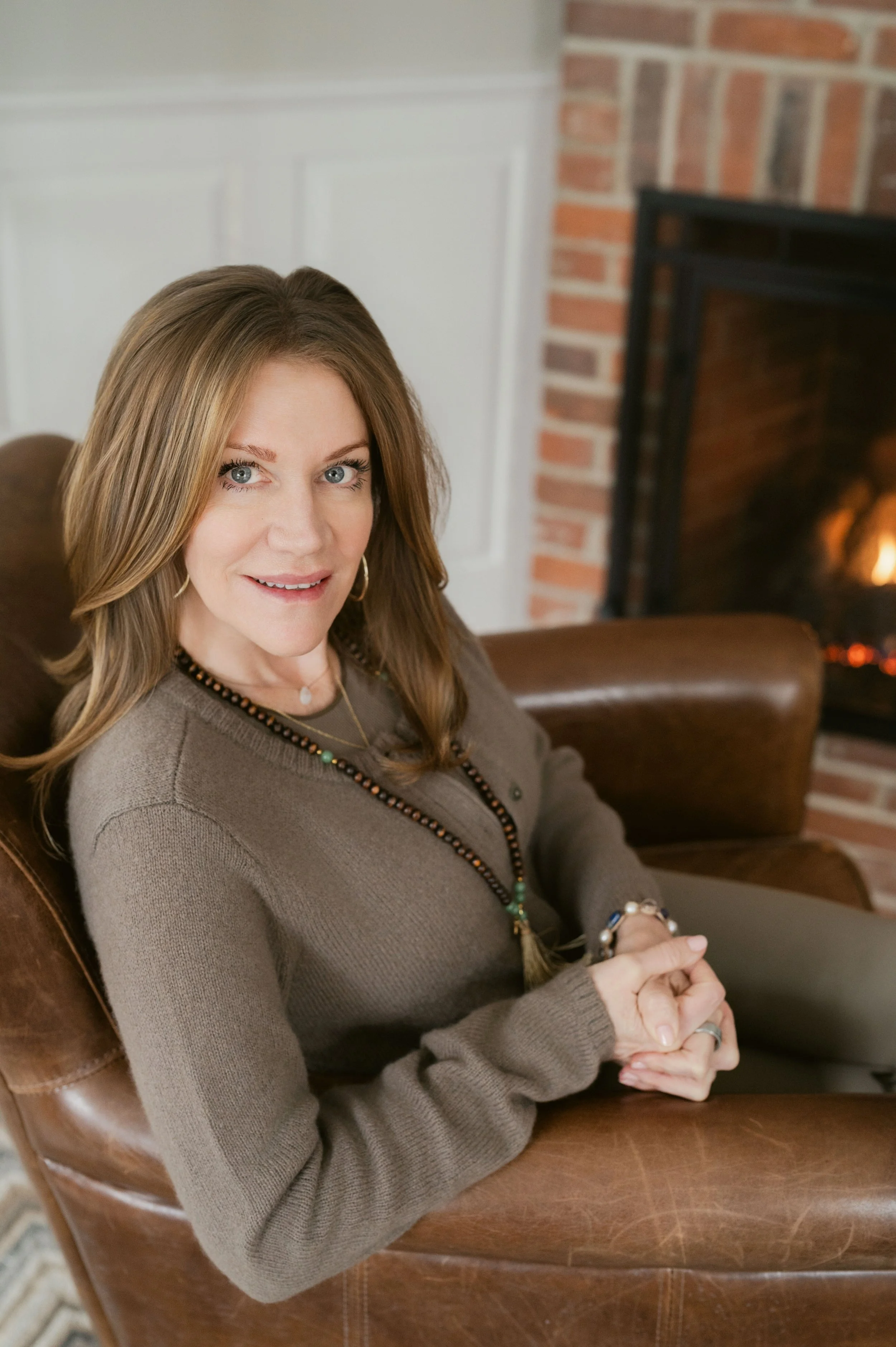 A woman with long, light brown hair and blue eyes sitting on a brown leather chair in a cozy room with a brick fireplace.