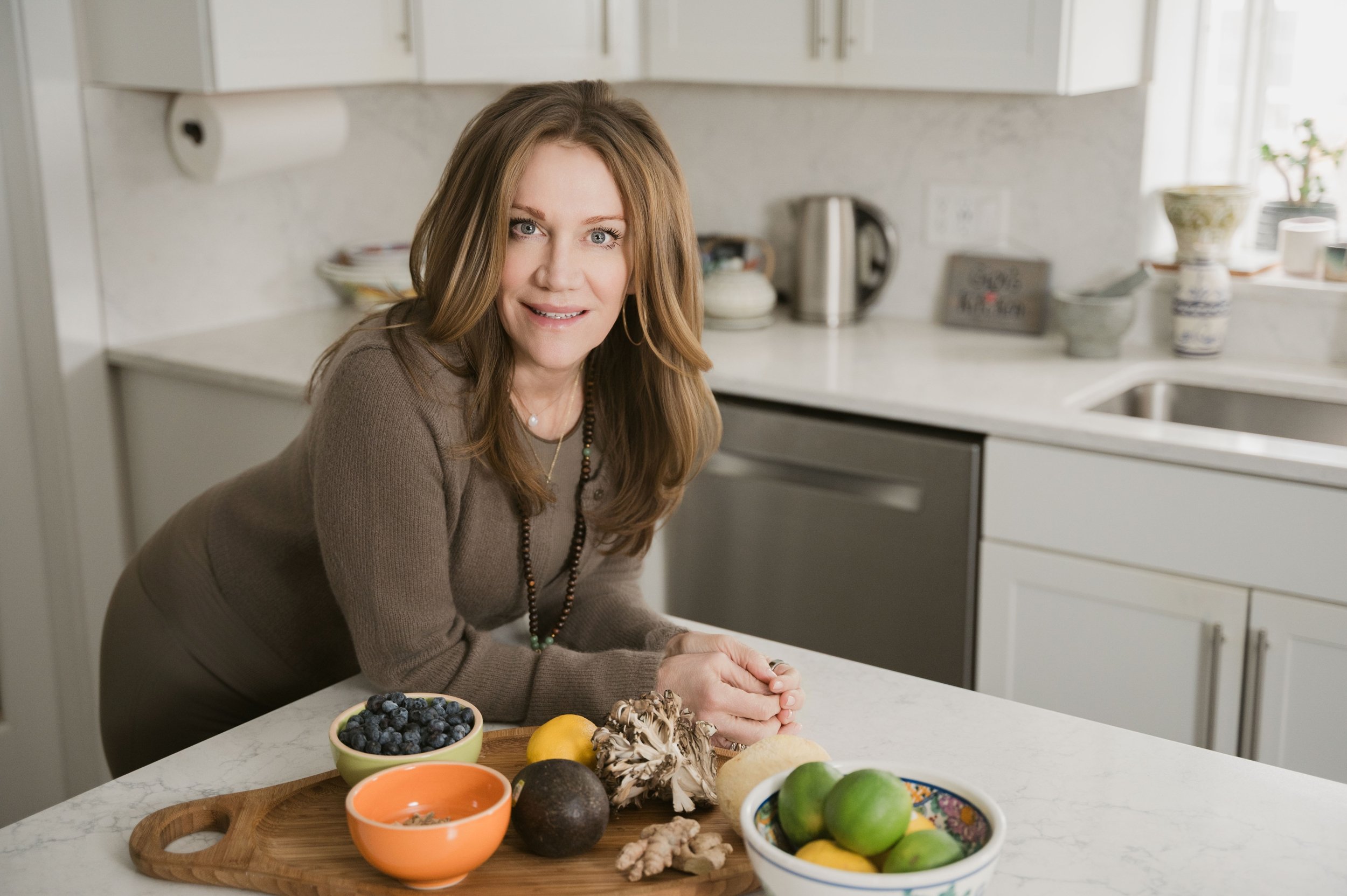 A woman with brown hair and blue eyes, wearing a brown sweater and jewelry, smiling at the camera while preparing food in a kitchen. There are bowls of blueberries, apples, ginger, and other ingredients on a wooden tray on the white marble kitchen counter.