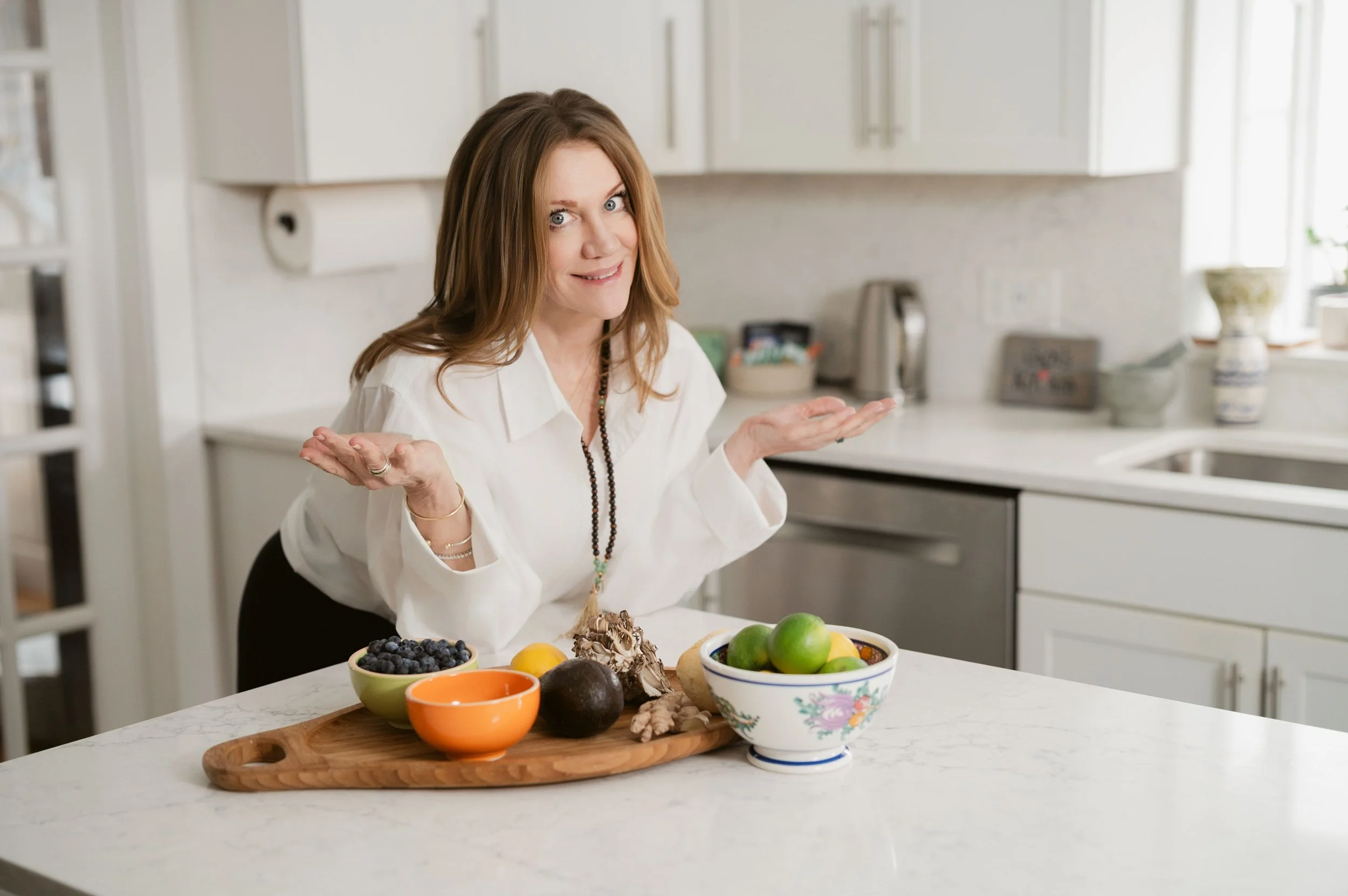 A woman with long brown hair and a white blouse sitting at a kitchen counter with a variety of fruits and dessert on a wooden tray, gesturing with a puzzled expression.