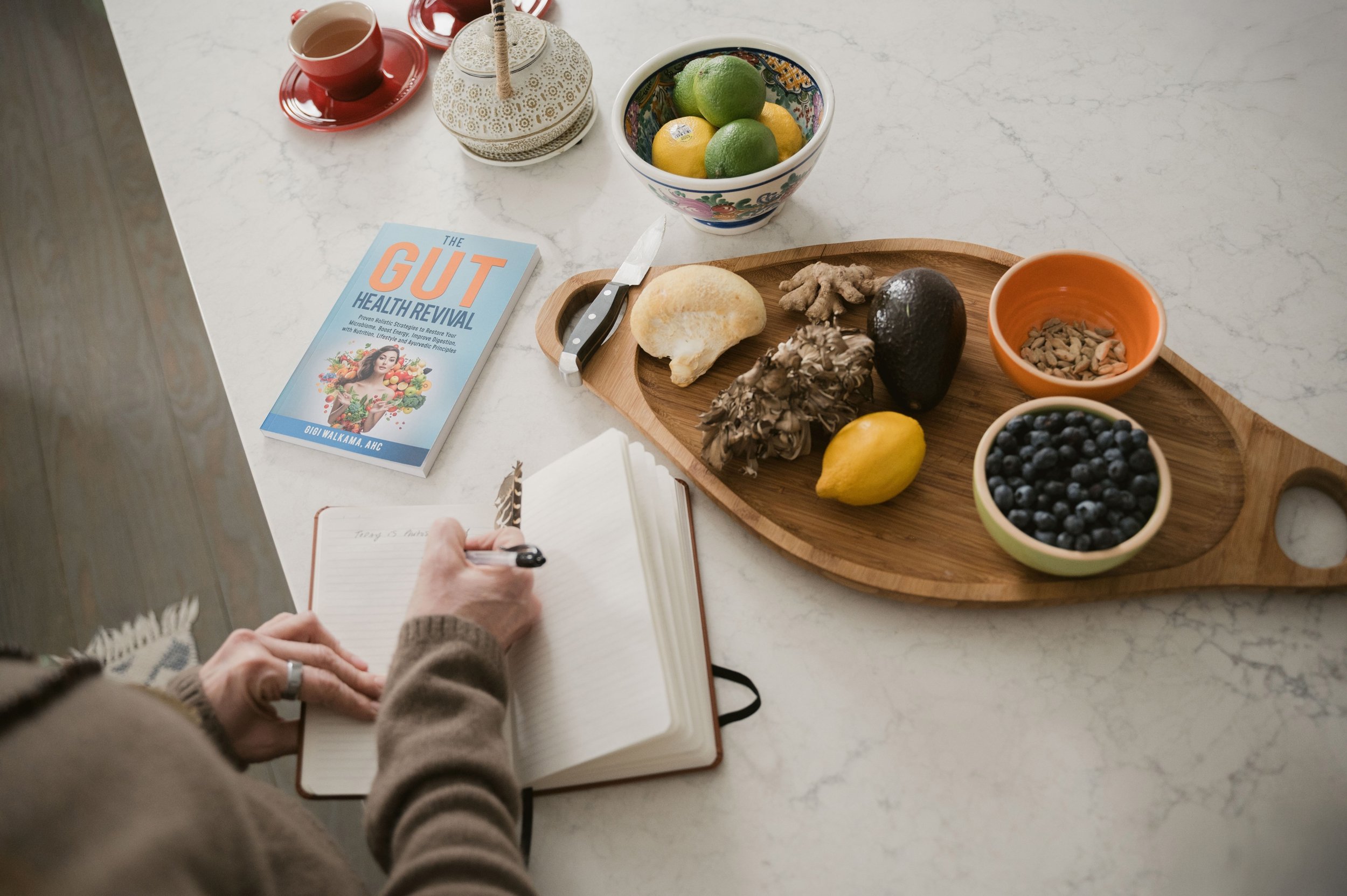 A person writing in a notebook at a table, with a book titled "The Gut Health Revival," a wooden tray with ginger, lemon, an avocado, a dark fruit, and sunflower seeds, and a bowl of blueberries, along with bowls of lemon and lime, an avocado, and a cup of tea.
