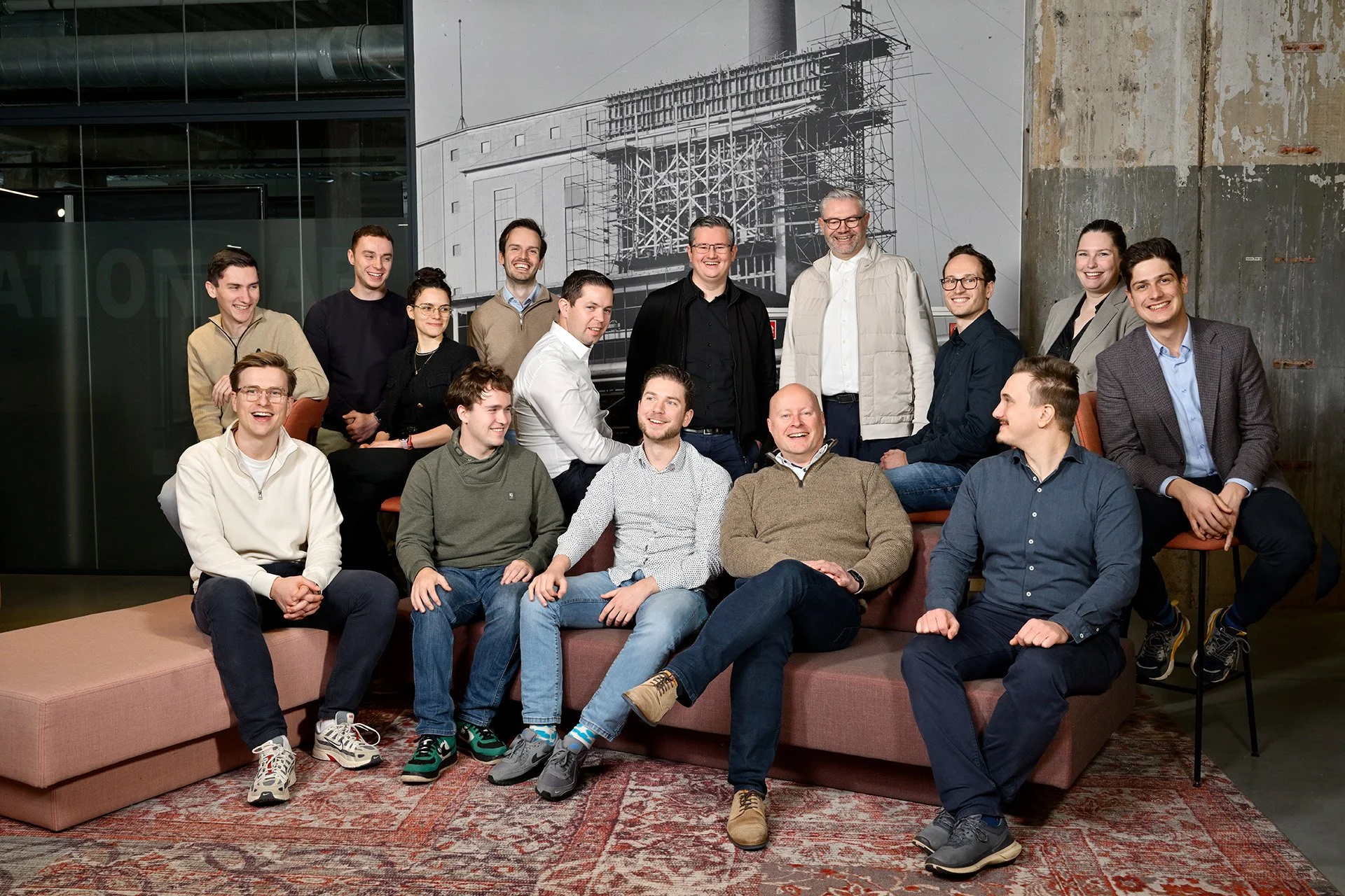 Group of diverse people smiling and sitting on a sofa and chairs in an industrial-style room with a large black-and-white photo of building scaffolding on the wall behind them.