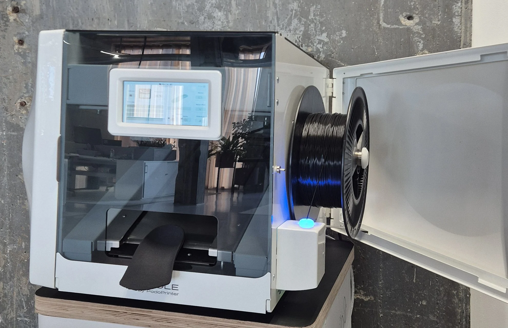 A white and transparent 3D printer with a black reel of filament on top, inside a clear enclosure, with a touchscreen control panel on the front.
