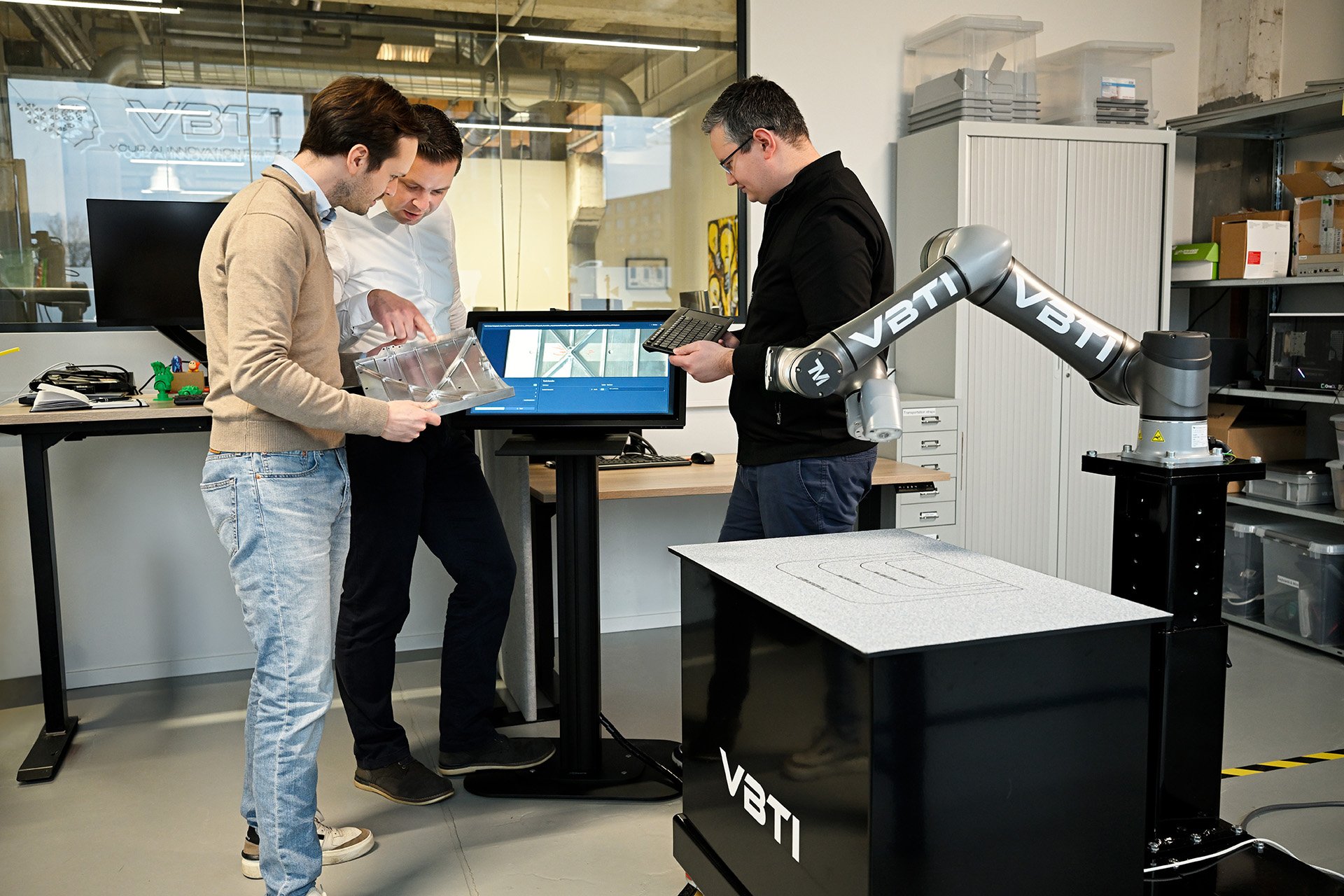 Three men are gathered around a table in a modern industrial lab or workshop, inspecting a clear, segmented device. One man is holding a keyboard, another is pointing at the device, and the third looks on. There is a large robotic arm labeled 'VBTI' attached to a stand near the table, and a monitor displaying a complex interface or blueprint. The background features shelves with boxes and containers, and a glass partition with more equipment visible behind it.