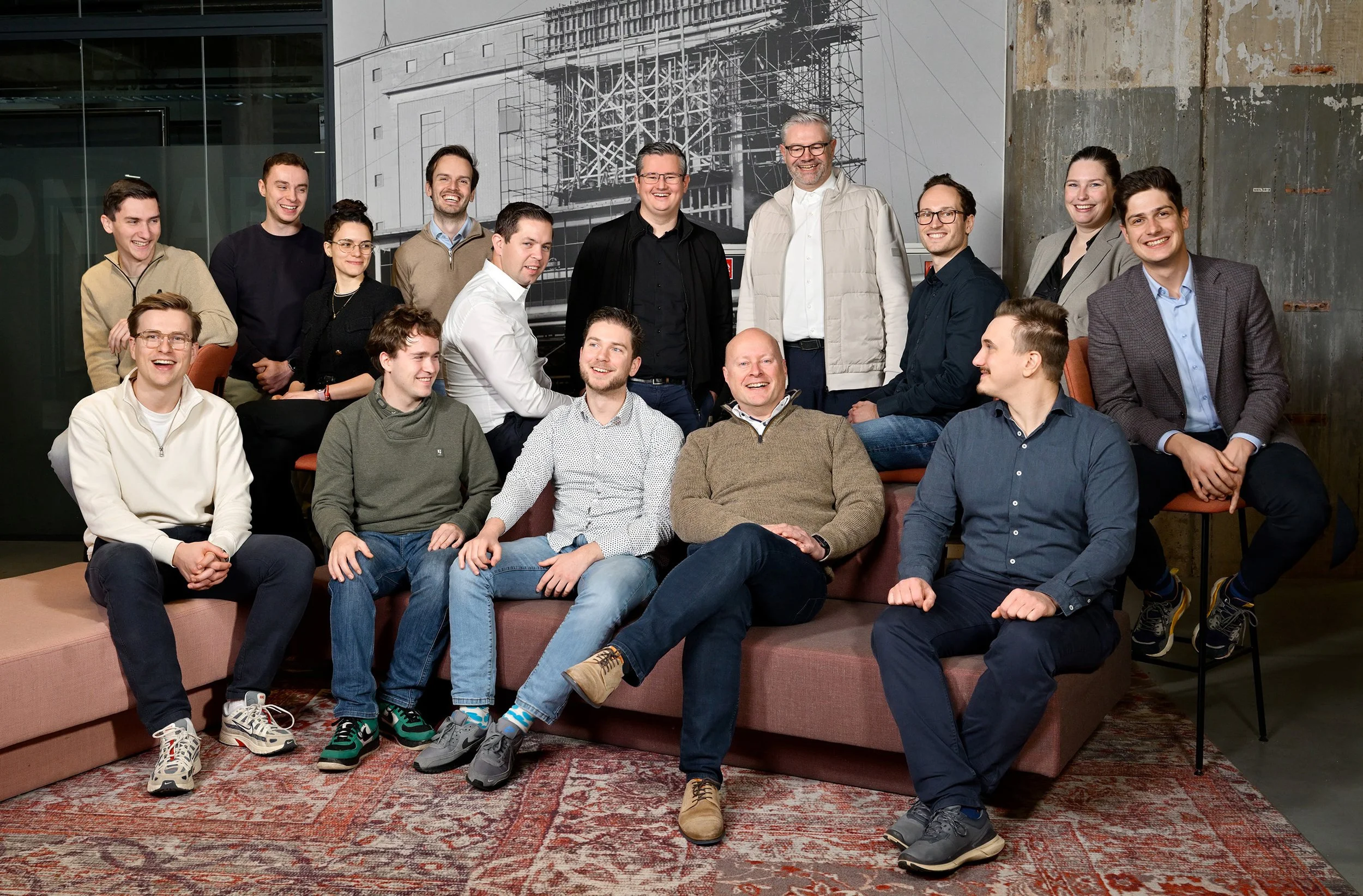 Group of sixteen people posing for a photo in an industrial-style room with a construction photo on the wall. They are sitting and standing on a patterned rug, smiling and dressed casually and business casual.