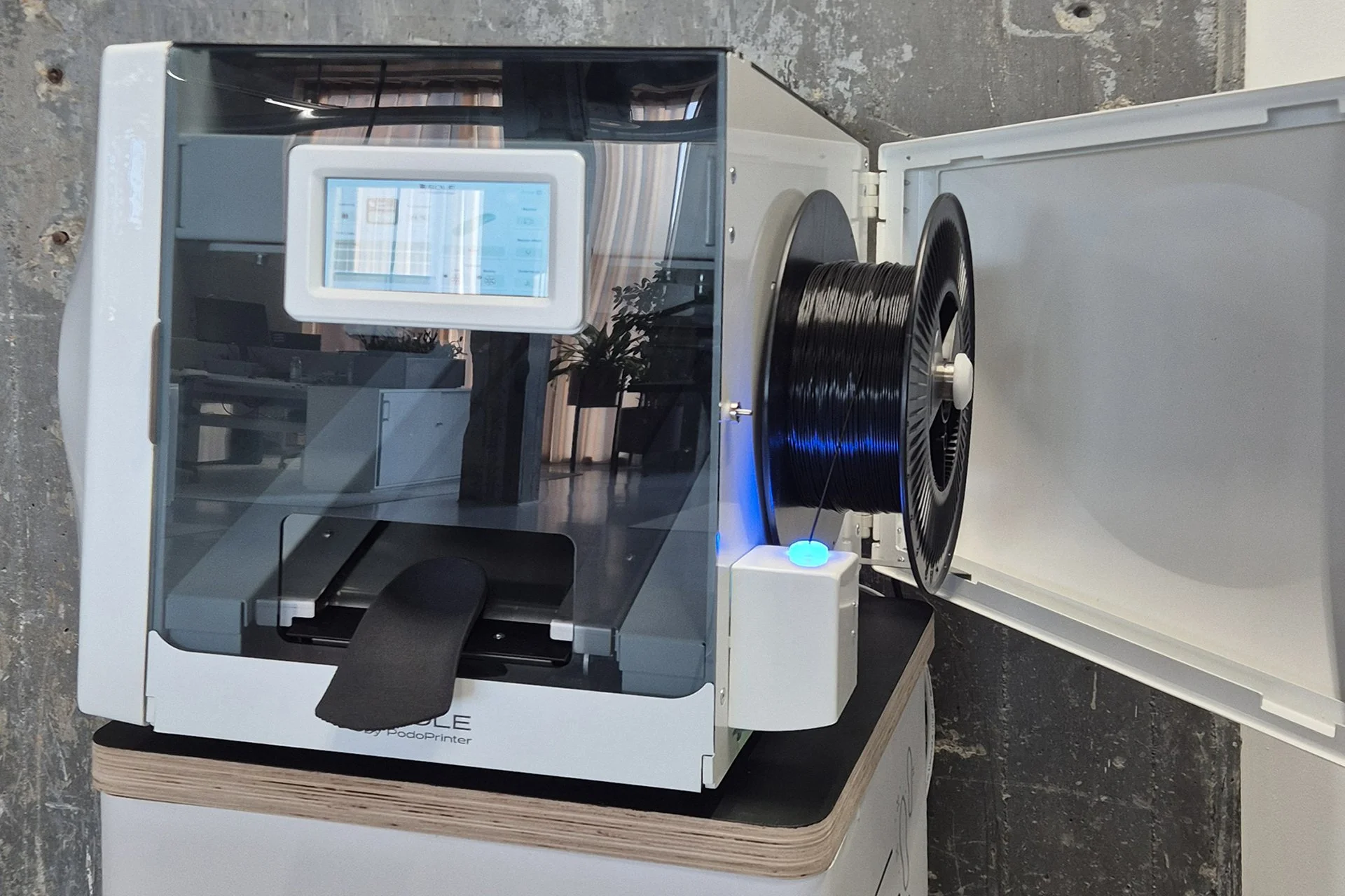 A 3D printer with a transparent cover positioned on a wooden table, with a spool of black filament on the side, in an indoor setting.