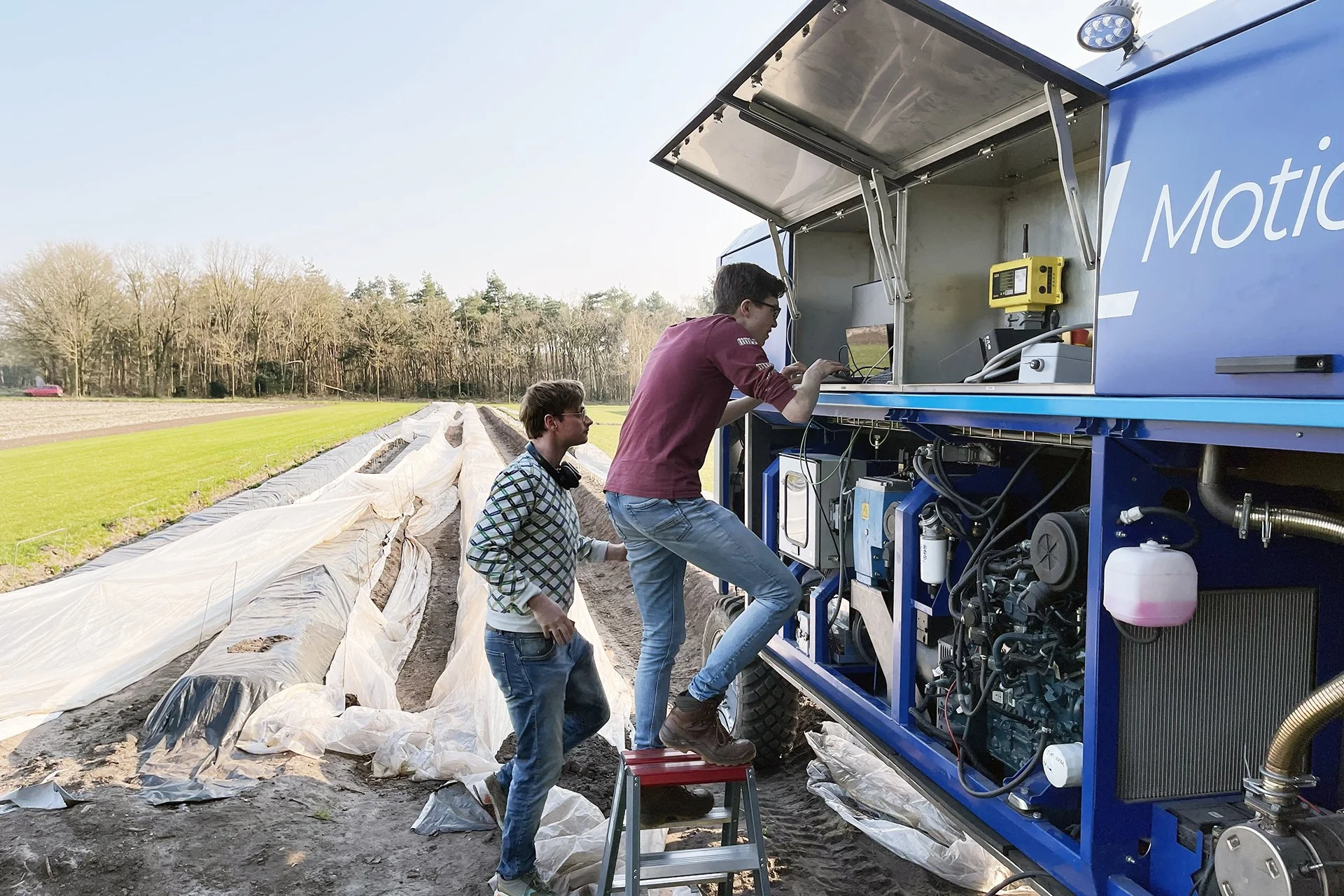 Two men are working with a large blue mobile technology unit in an agricultural field, with plastic-covered rows of plants and trees in the background.