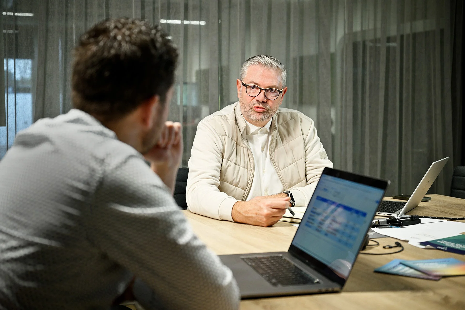 Two men attending a meeting in an office with laptops, papers, and office supplies on the table, one man facing the camera with glasses and gray hair, engaging in conversation.