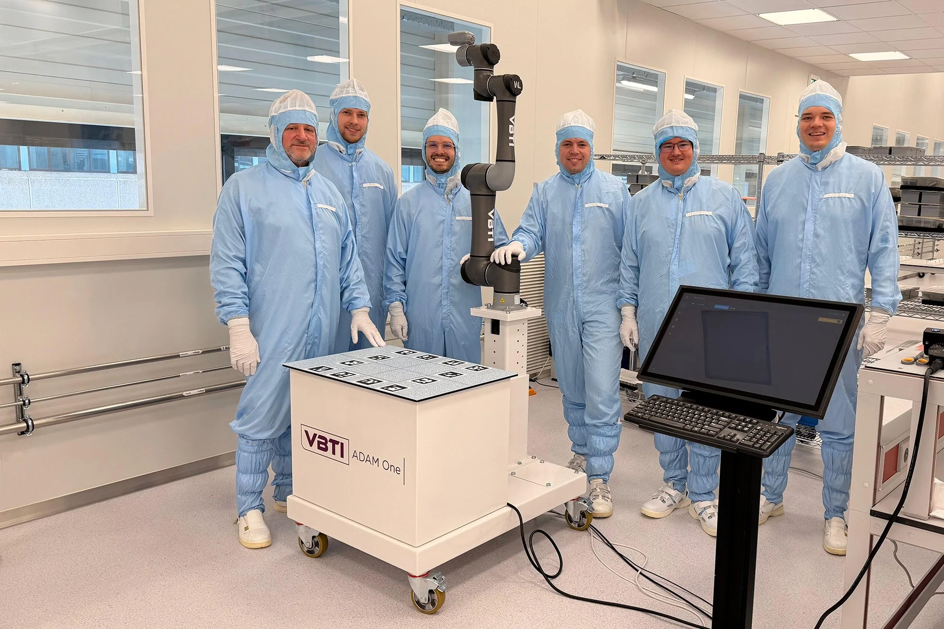 A team of six scientists in blue cleanroom suits and hairnets posing in a laboratory with robotic equipment and a computer. They are standing behind a white cart labeled 'VBTI ADAM One' with electronic components, and a robotic arm is positioned between them.