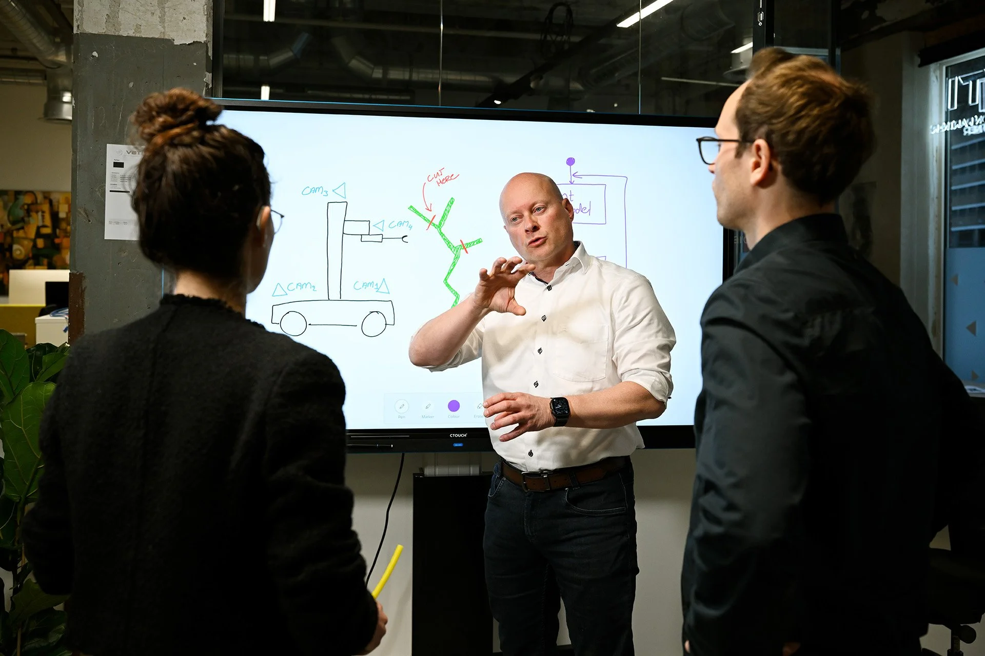 Man in white shirt explaining a diagram on a large digital screen to two women in black attire, in an industrial-style office space with plants and reflective ceiling panels.