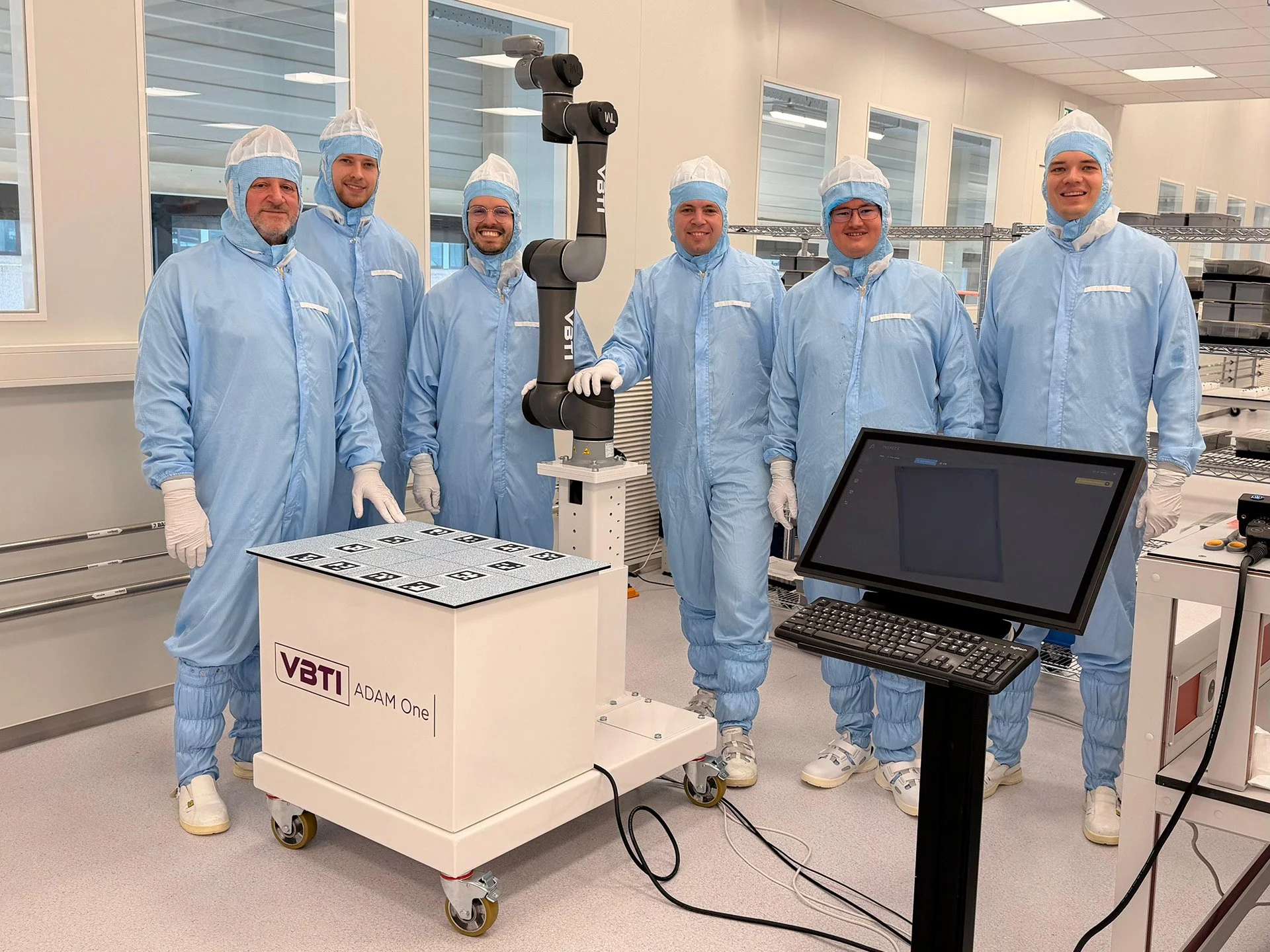 Six people in blue cleanroom suits and hair covers standing in a laboratory. They are smiling and standing around a robotic arm and a small white table with the label 'VBTI ADAM One'. There are shelves and windows in the background.