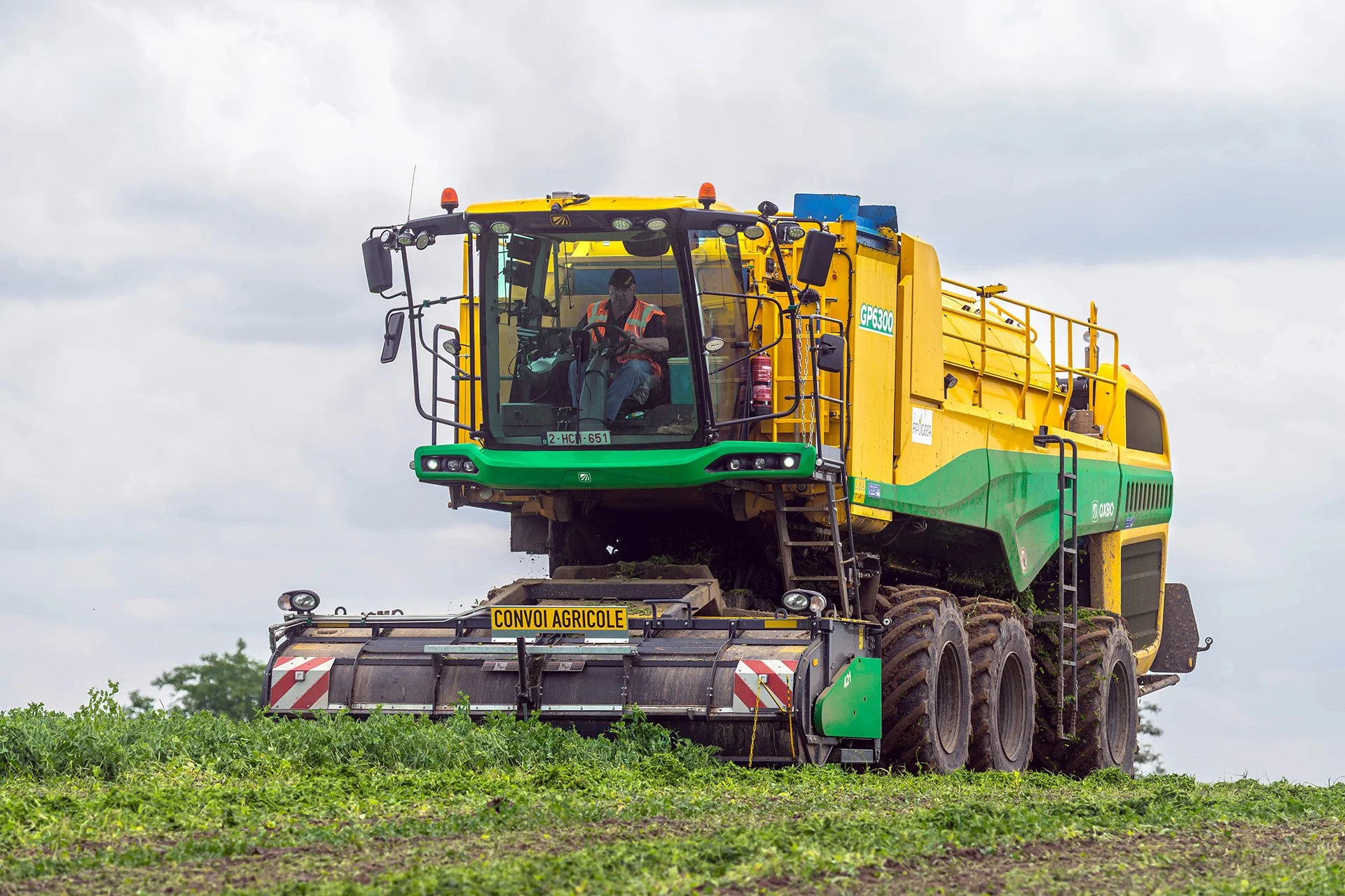A large yellow and green agricultural machine with a driver inside, working in a field under cloudy skies.