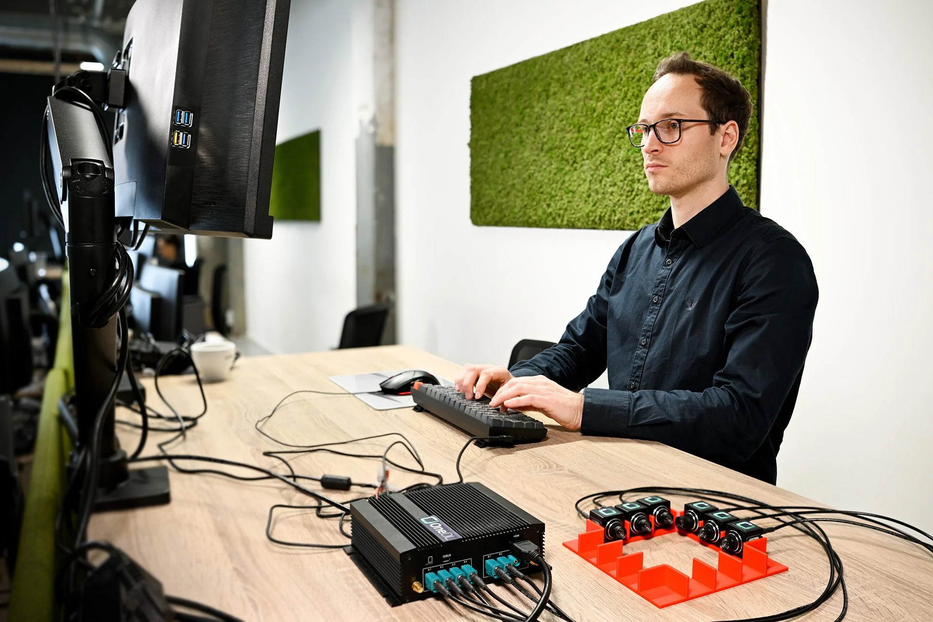 A man wearing glasses working on a computer with various electronic devices and connectors on a wooden desk in an office setting.