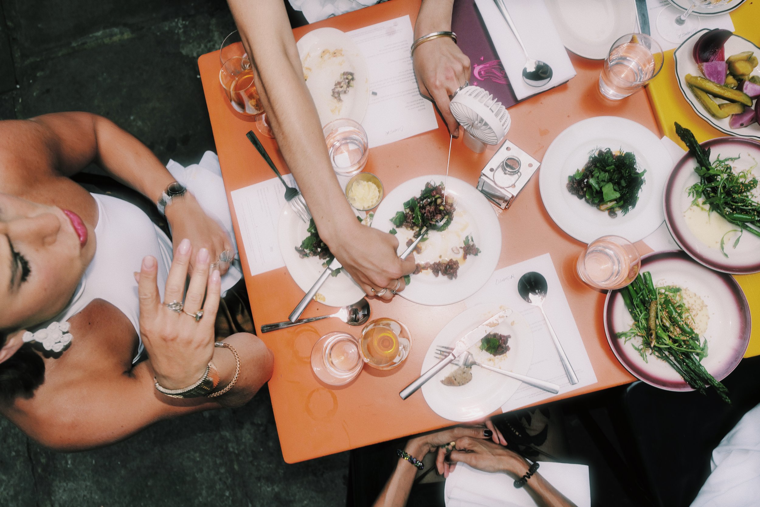 Overhead view of a group of people sitting at a table with various plates of food, glasses of water, and utensils, during a meal.